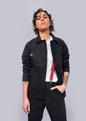 S=Wearing The Essential Utility Jacket and comfortable stretch pants, a person with short curly hair pairs their look with a white shirt and red suspenders, standing confidently against a white background.