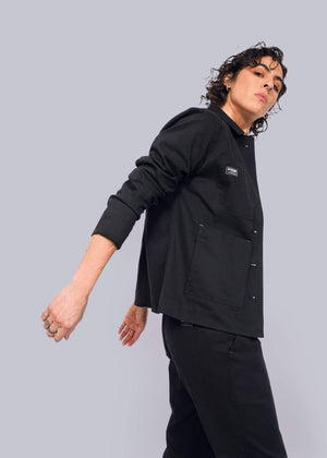 S=With curly hair and dressed in black, a person poses against a white background wearing The Essential Utility Jacket, featuring deep pockets and stretch comfort. Their head is slightly tilted as they gaze to the side with one arm bent.