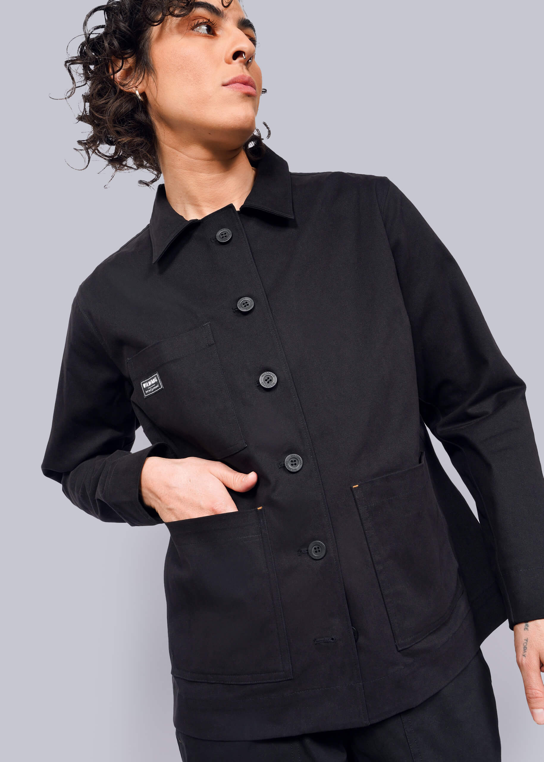 S=Wearing The Essential Utility Jacket in black, they stand against a plain background with their left hand in a deep front pocket. A small square label adorns the chest as their curly hair enhances the effortless blend of style and stretch comfort, while they gaze off to the side.