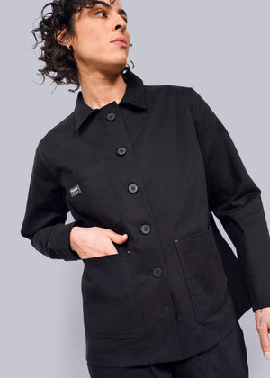S=Wearing The Essential Utility Jacket in black, they stand against a plain background with their left hand in a deep front pocket. A small square label adorns the chest as their curly hair enhances the effortless blend of style and stretch comfort, while they gaze off to the side.