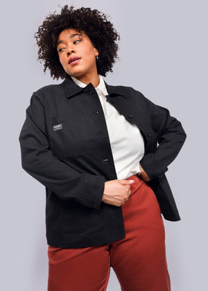 1X=A person with curly hair models The Essential Utility Jacket, featuring deep pockets, over a white shirt and red pants. They pose with one hand on their hip, looking to the side against a plain white background.