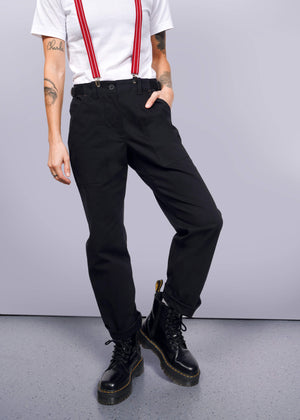 S=The person embodies a workwear style in black Essential Utility Pants paired with red suspenders and solid black boots. Against a plain background, their right arm rests on their hip, showcasing tattoos on both arms, while the other hand casually hangs by their side.