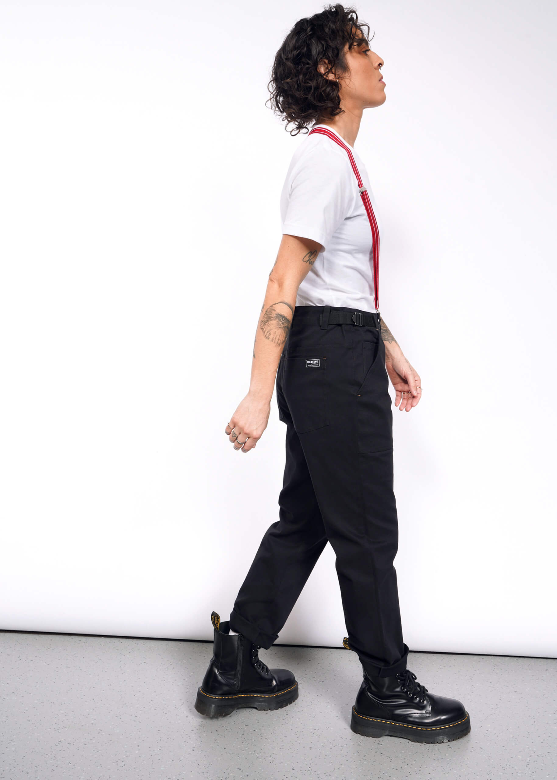 S=Striding confidently against a white background, a person with curly hair sports The Essential Utility Pant in black with red suspenders, paired with a white t-shirt. Their outfit blends stretch cotton comfort and workwear style, completed by black boots and tattoos visible on their arms.