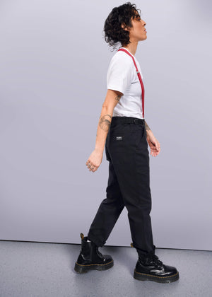 S=Striding confidently against a white background, a person with curly hair sports The Essential Utility Pant in black with red suspenders, paired with a white t-shirt. Their outfit blends stretch cotton comfort and workwear style, completed by black boots and tattoos visible on their arms.