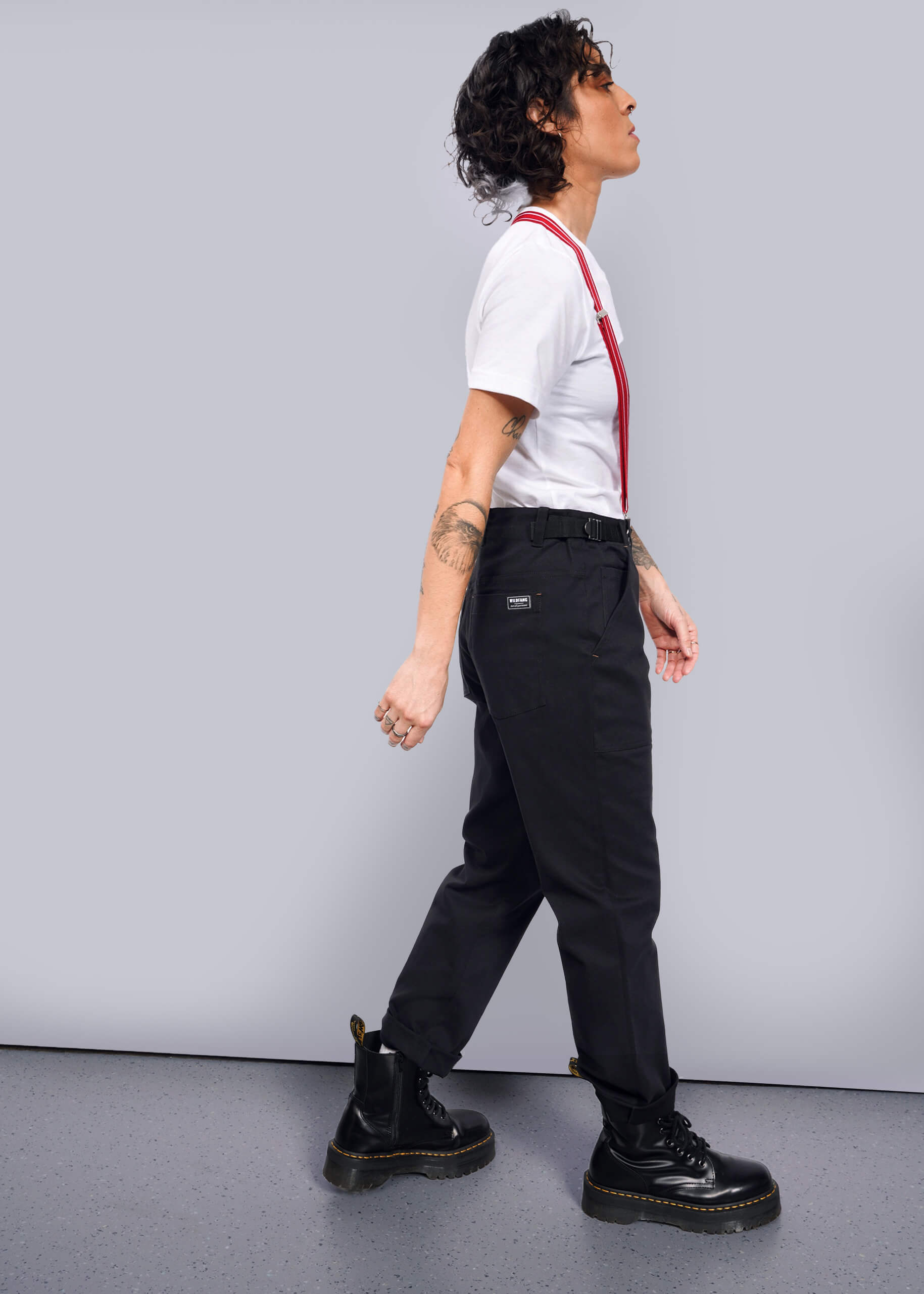 S=Striding confidently against a white background, a person with curly hair sports The Essential Utility Pant in black with red suspenders, paired with a white t-shirt. Their outfit blends stretch cotton comfort and workwear style, completed by black boots and tattoos visible on their arms.