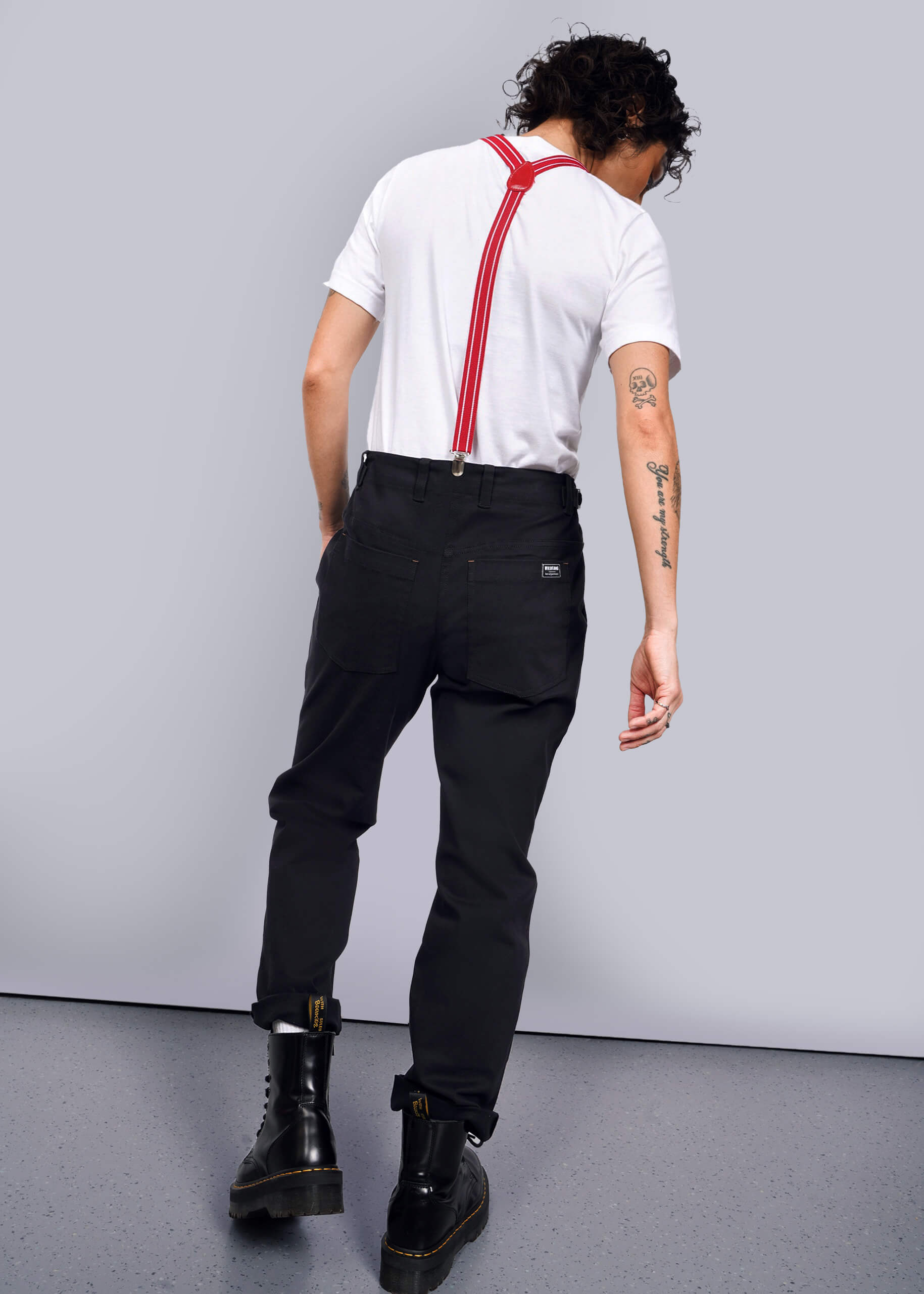 S=Facing away, the person wears a white t-shirt and black boots. Curly hair complements red suspenders over The Essential Utility Pant for a workwear look. Tattoos on their left arm add personal flair to the stretch cotton ensemble.