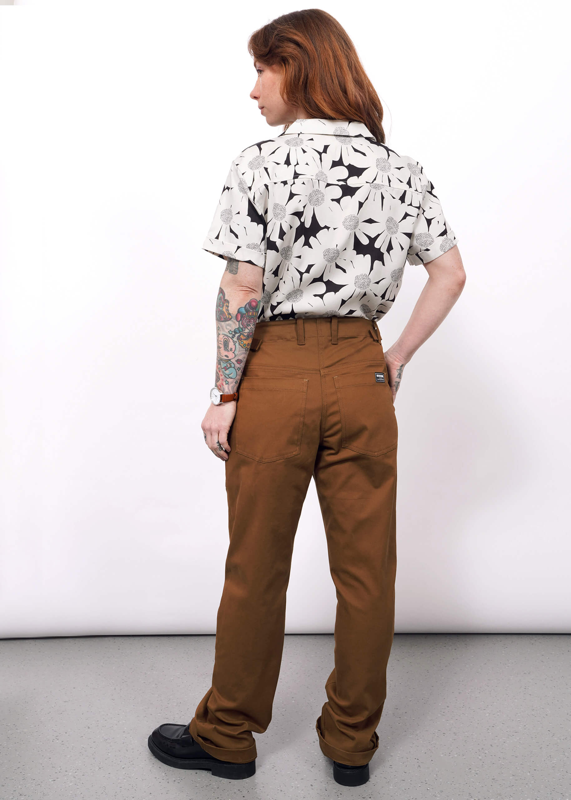 S=A person with long brown hair stands facing away, wearing The Essential Utility Pant in brown, featuring spacious pockets, paired with a black and white floral short-sleeve shirt and black shoes. A colorful tattoo shows on their left forearm against a plain white background.