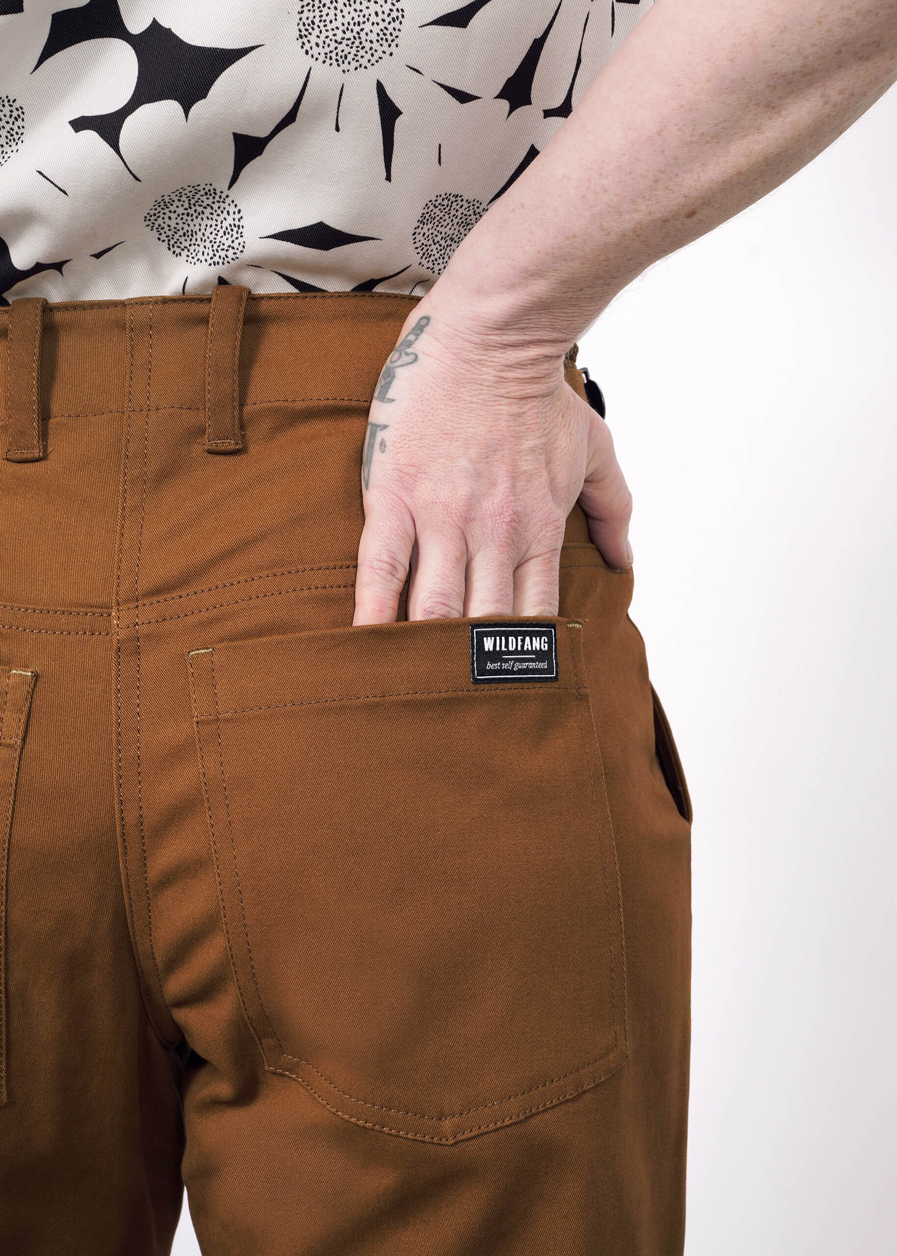S=A person in The Essential Utility Pant by Wildfang, rust-colored with spacious back pockets, slips their tattooed hand inside. They wear a white shirt with a black abstract floral pattern.