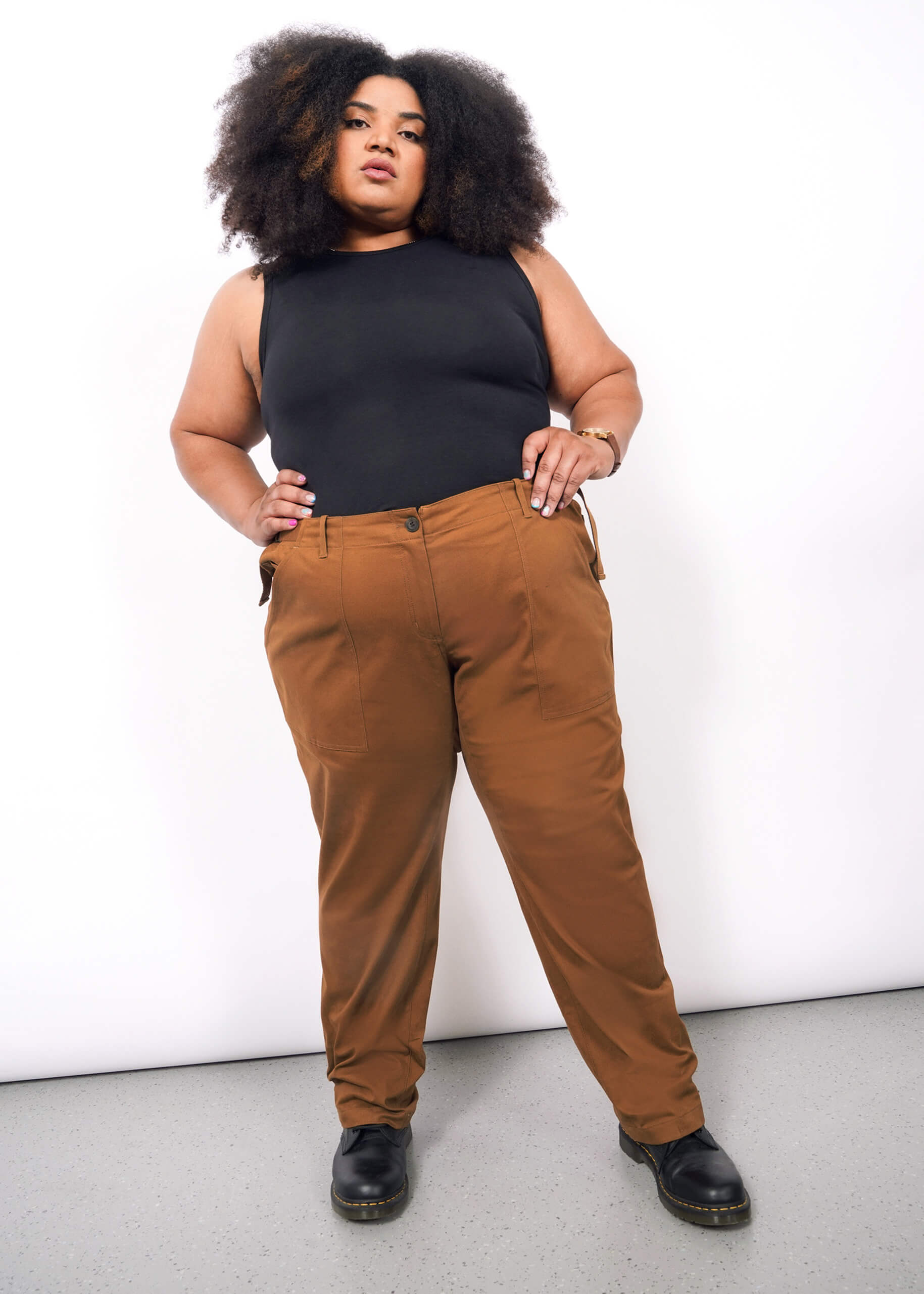 1X=A person with voluminous curly hair wears a sleeveless black top, The Essential Utility Pant in rust with spacious pockets, and black shoes, standing confidently against a plain white background.