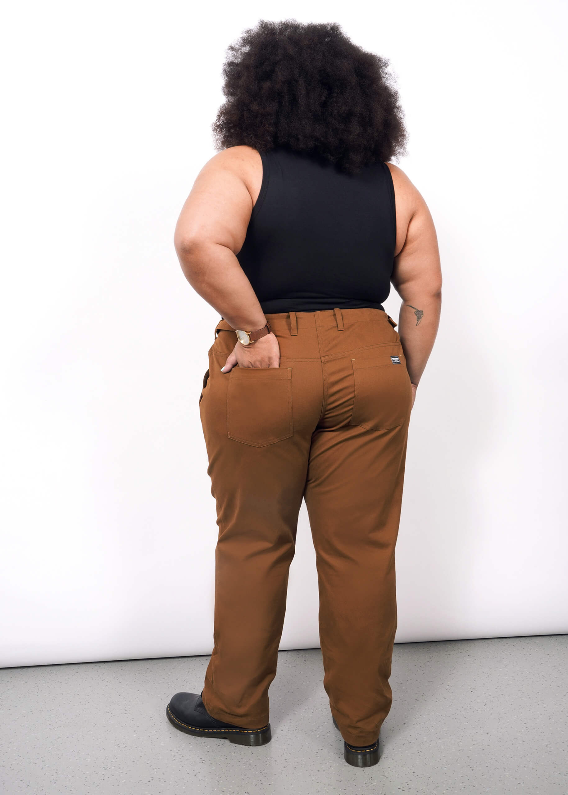 1X=Wearing The Essential Utility Pant, a person with curly hair and a black sleeveless top stands facing away from the camera, one hand in their back pocket.
