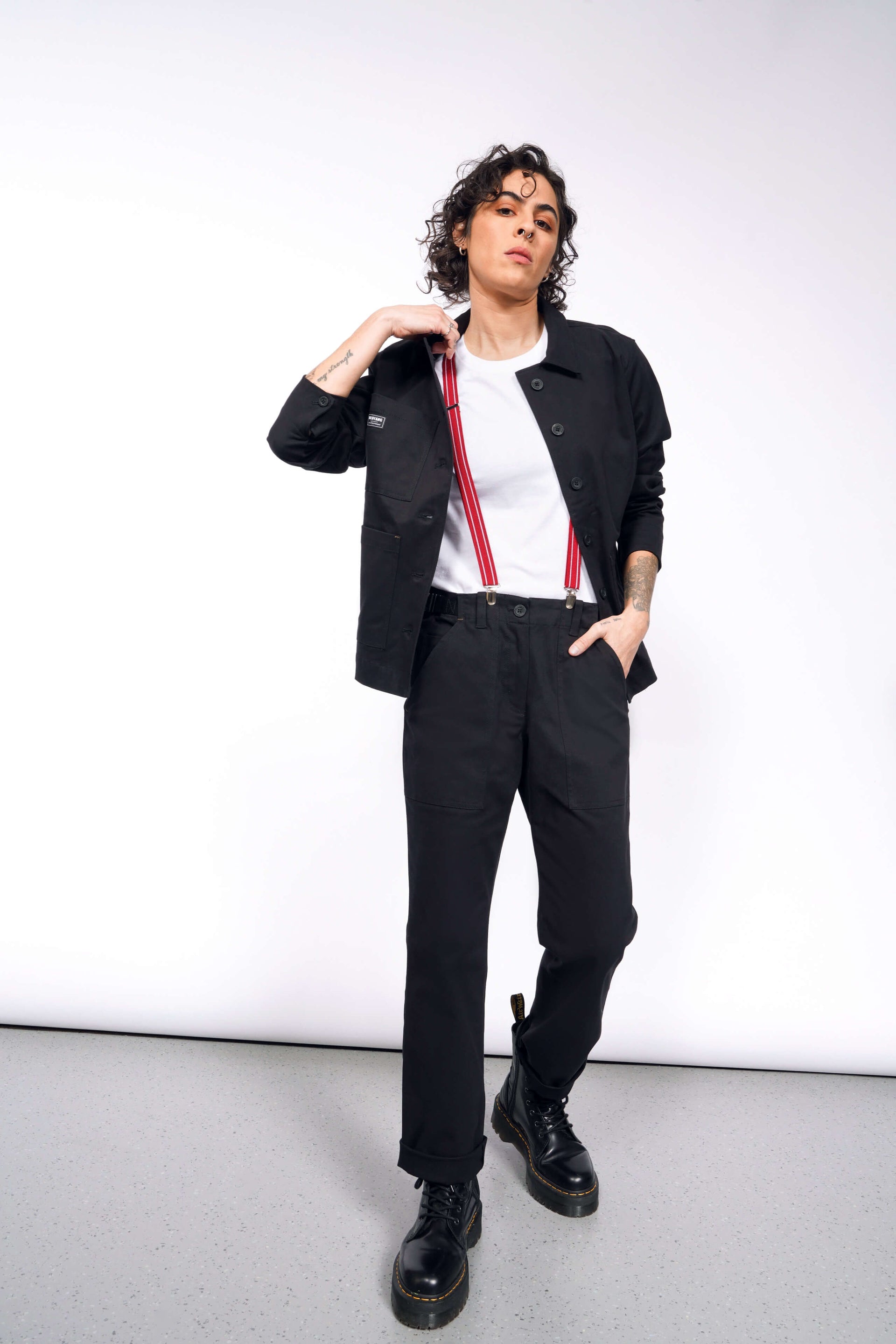 S=A person with curly hair stands confidently in a black jacket, The Essential Utility Pant, and black boots. They sport red suspenders over a stretch cotton white t-shirt, with one hand in their pocket against a simple light-colored background, embodying the workwear style.