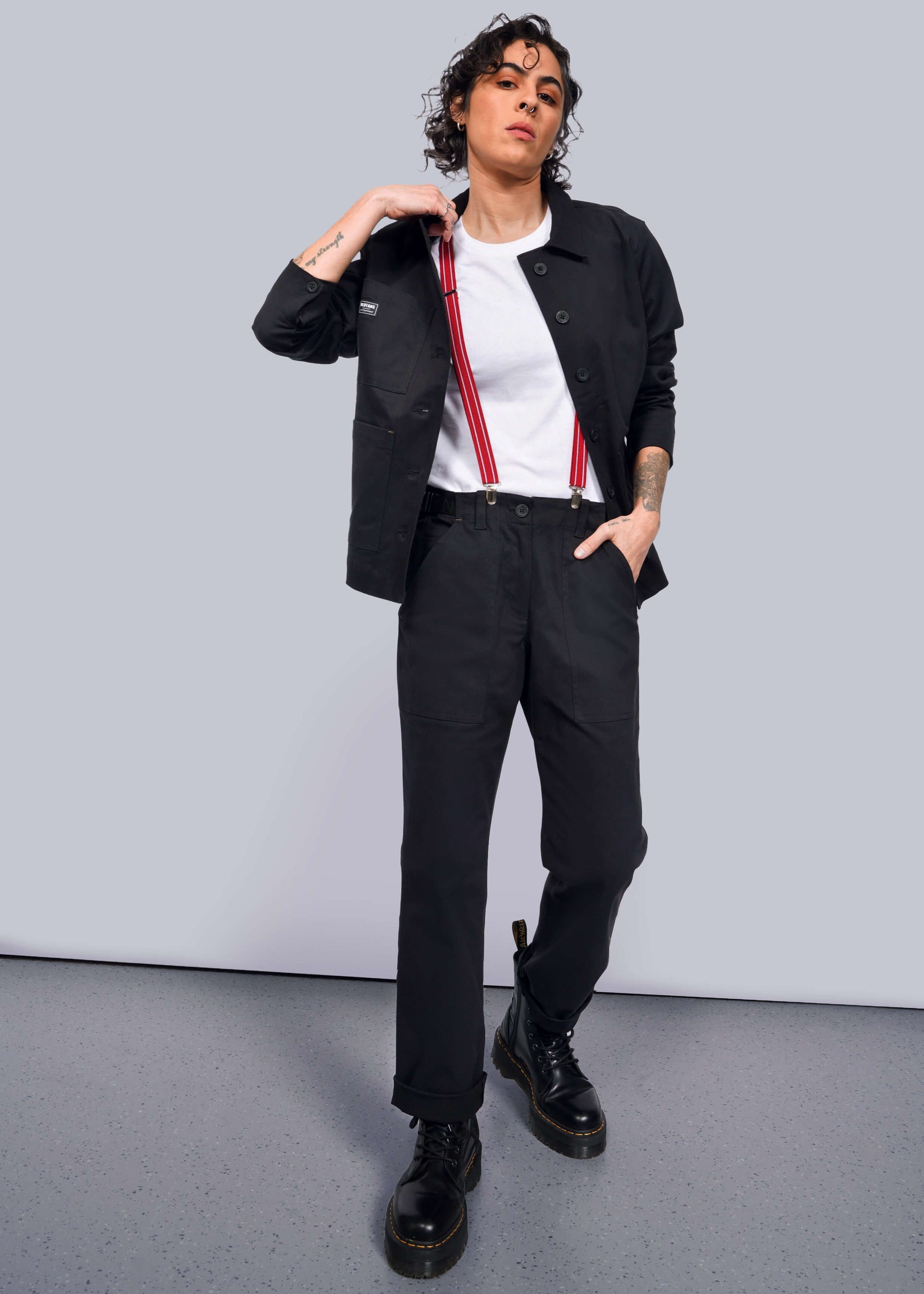 S=A person with curly hair stands confidently in a black jacket, The Essential Utility Pant, and black boots. They sport red suspenders over a stretch cotton white t-shirt, with one hand in their pocket against a simple light-colored background, embodying the workwear style.