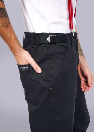 S=Someone wearing The Essential Utility Pant in black, featuring a sleek belt and bold red suspenders. A hand casually rests in the pocket, showcasing the label. The stretch cotton fabric pairs with a crisp white shirt for a seamless modern workwear look.