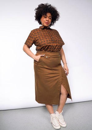 1X= A person with curly hair poses confidently in a patterned brown shirt and The Essential Utility Wrap Skirt, featuring a zippered opening and adjustable waist buckle at the front. They are wearing white high-top sneakers, standing against a plain gray background.