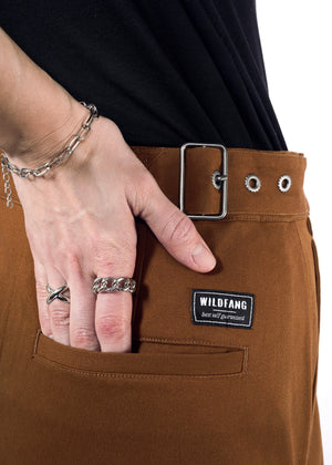 S=Close-up of a person in rust-colored pants featuring an adjustable waist buckle and rectangular silver clasp. A hand with silver rings and a chain bracelet rests in the back pocket, showing Wildfang branding. The outfit is completed with a sleek black shirt. Product: The Essential Utility Wrap Skirt.
.