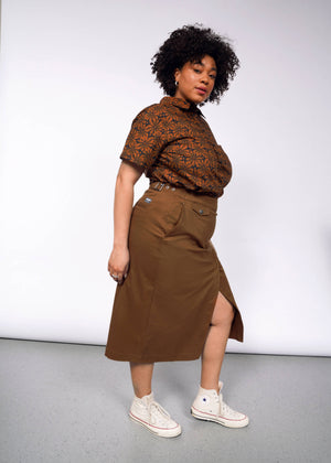 1X=A person with curly hair wears a patterned brown shirt and The Essential Utility Wrap Skirt, styled with an adjustable waist buckle, standing confidently against a plain backdrop. They complete the look with white high-top sneakers.