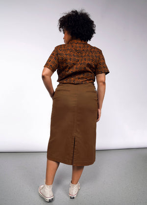 1X= A person with curly hair stands with their back to the camera, wearing a brown geometric-patterned shirt and The Essential Utility Wrap Skirt, which has an adjustable waist buckle. They are in white sneakers on a gray floor against a white backdrop.