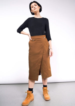 S= A person dressed in a black long-sleeve top, The Essential Utility Wrap Skirt with button details and an adjustable waist buckle, complemented by tan ankle boots. Their short hair is styled as they confidently pose with one hand on their hip in a minimalist space.