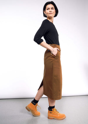 S=A person with dark hair is wearing a black shirt, The Essential Utility Wrap Skirt in brown with a zippered opening, and mustard yellow boots. They stand in profile against a plain white background, hands in pockets, maintaining a neutral expression.