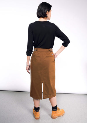 S=A person with short black hair wears a black long-sleeve top and The Essential Utility Wrap Skirt, a brown midi with back slit and adjustable waist buckle. Facing away from the camera, they sport tan boots on a light gray floor against a white wall.