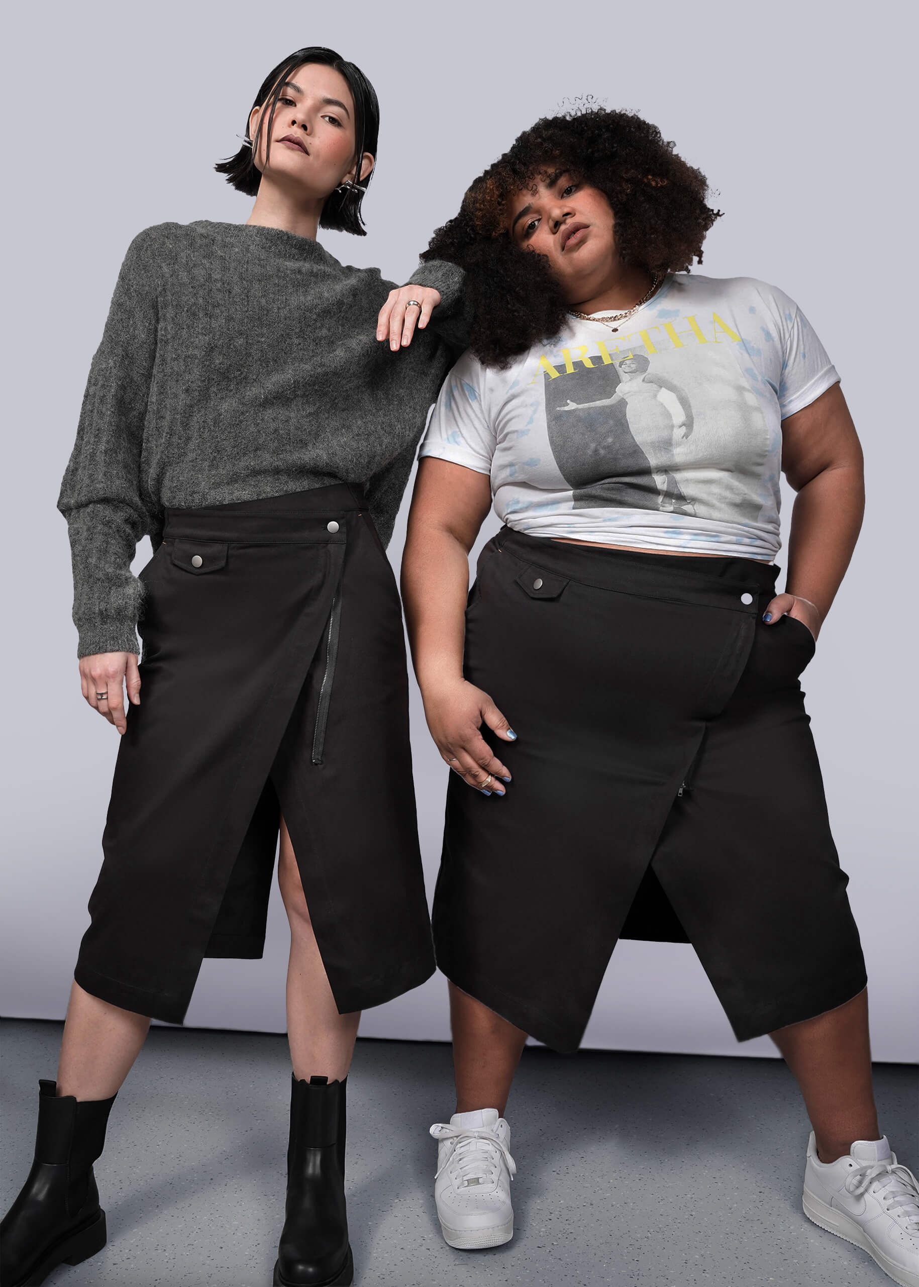 Two women showcase The Essential Utility Wrap Skirt, styled with a gray sweater and beanie or a graphic tee and sneakers. Each pairs the black midi skirt’s front slit with unique looks, standing against a plain background.
