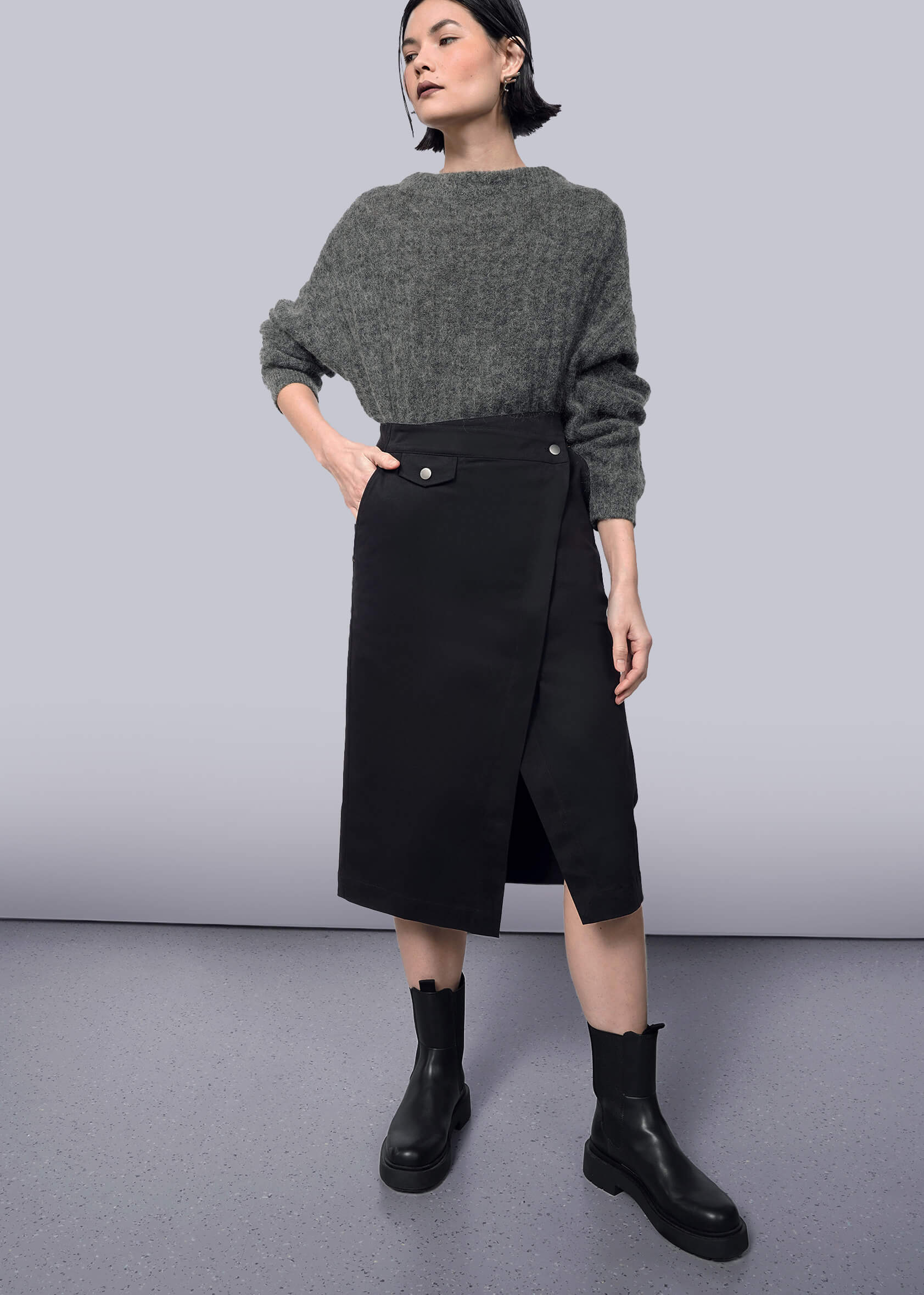 S= A woman poses against a white backdrop in a gray sweater and The Essential Utility Wrap Skirt—black, midi length, with buttons and an asymmetric waist. She pairs the look with black ankle boots and styles her short dark hair confidently.