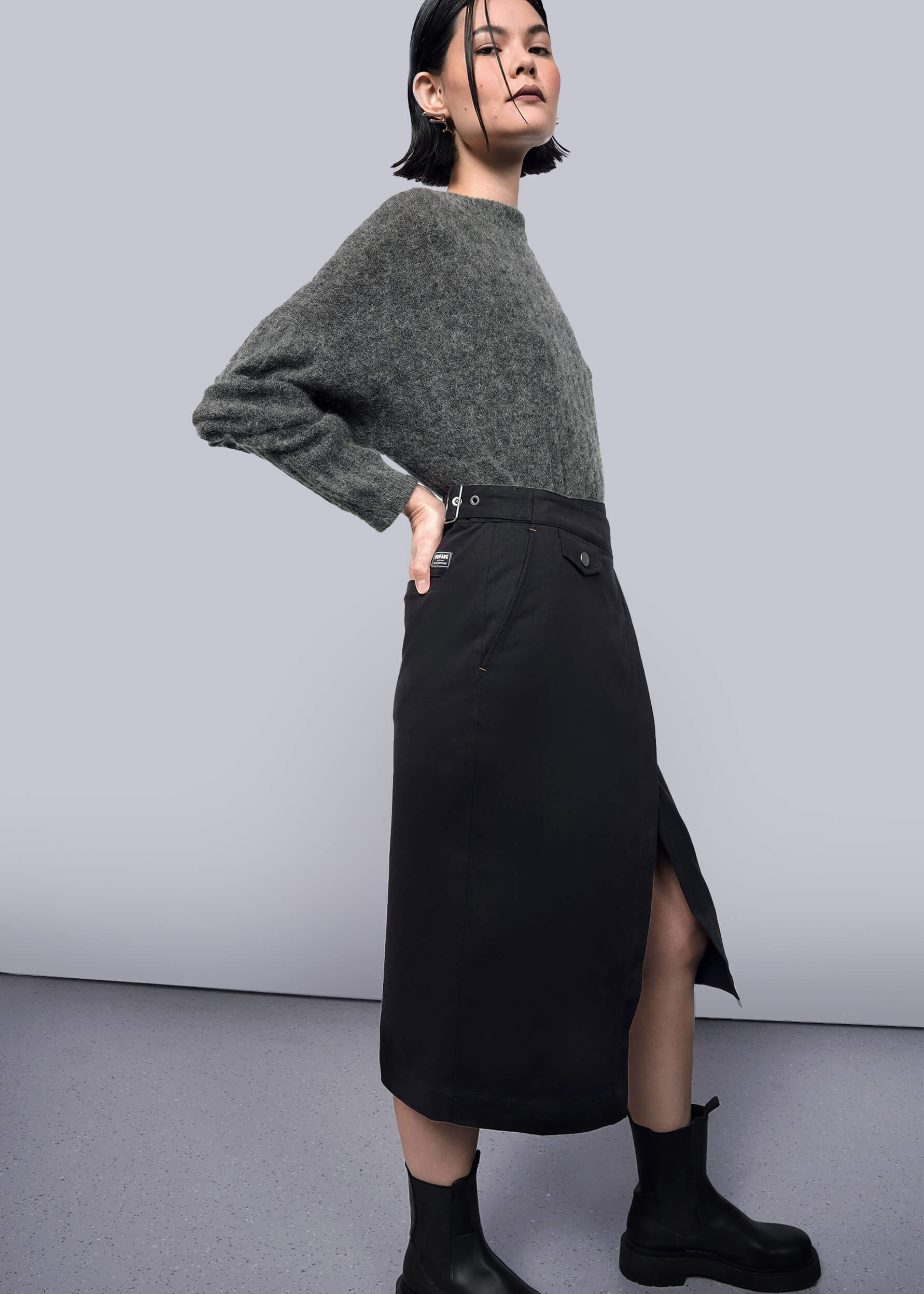 S= A woman stands sideways against a white background, confidently posing with one hand on her hip in a gray fuzzy sweater, black ankle boots, and The Essential Utility Wrap Skirt featuring an asymmetrical waist detail.