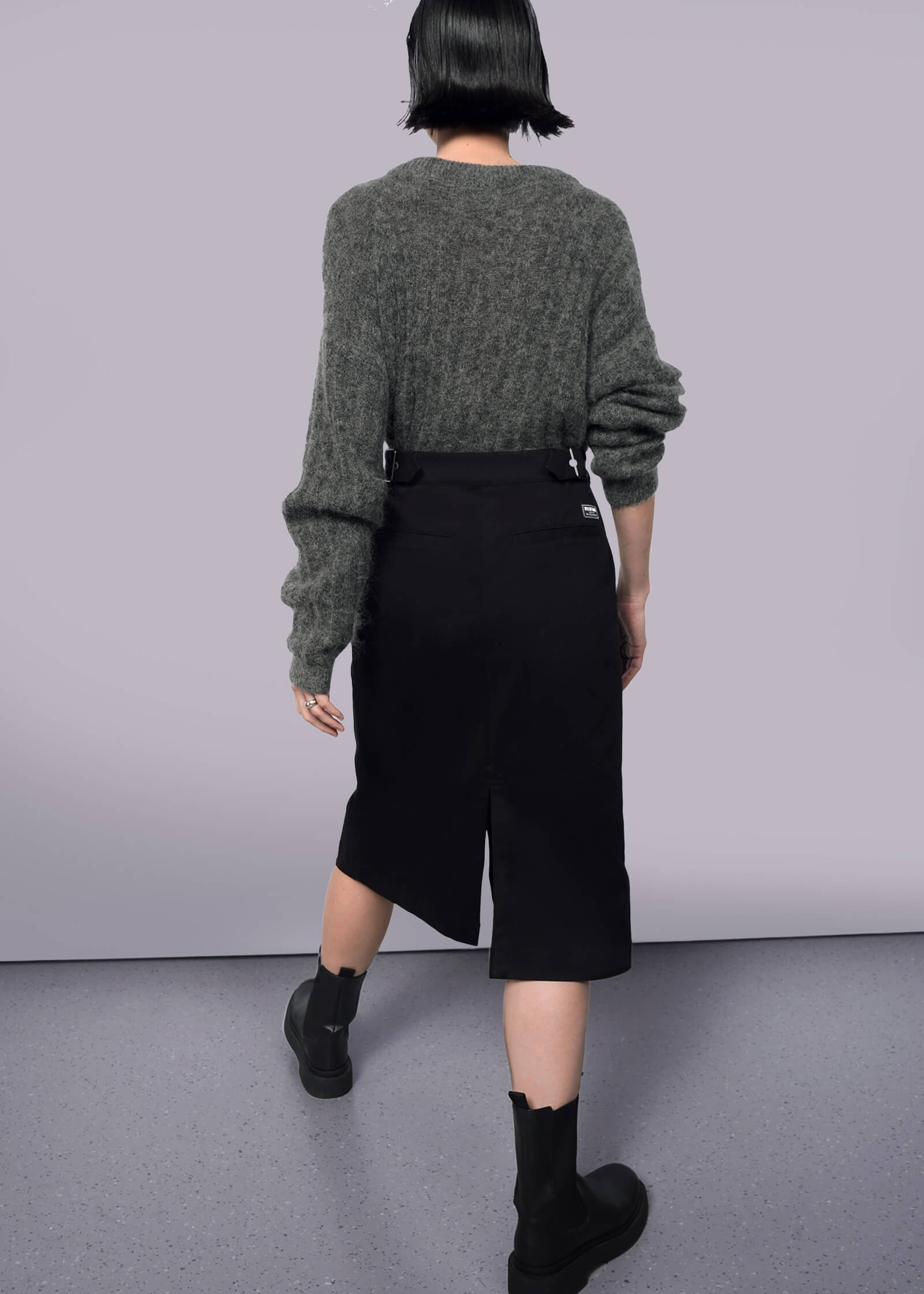S= A person with short dark hair faces away, wearing a loose gray sweater with The Essential Utility Wrap Skirt—black, midi-length with back slit and adjustable waistband—and black ankle boots, against a plain white background.