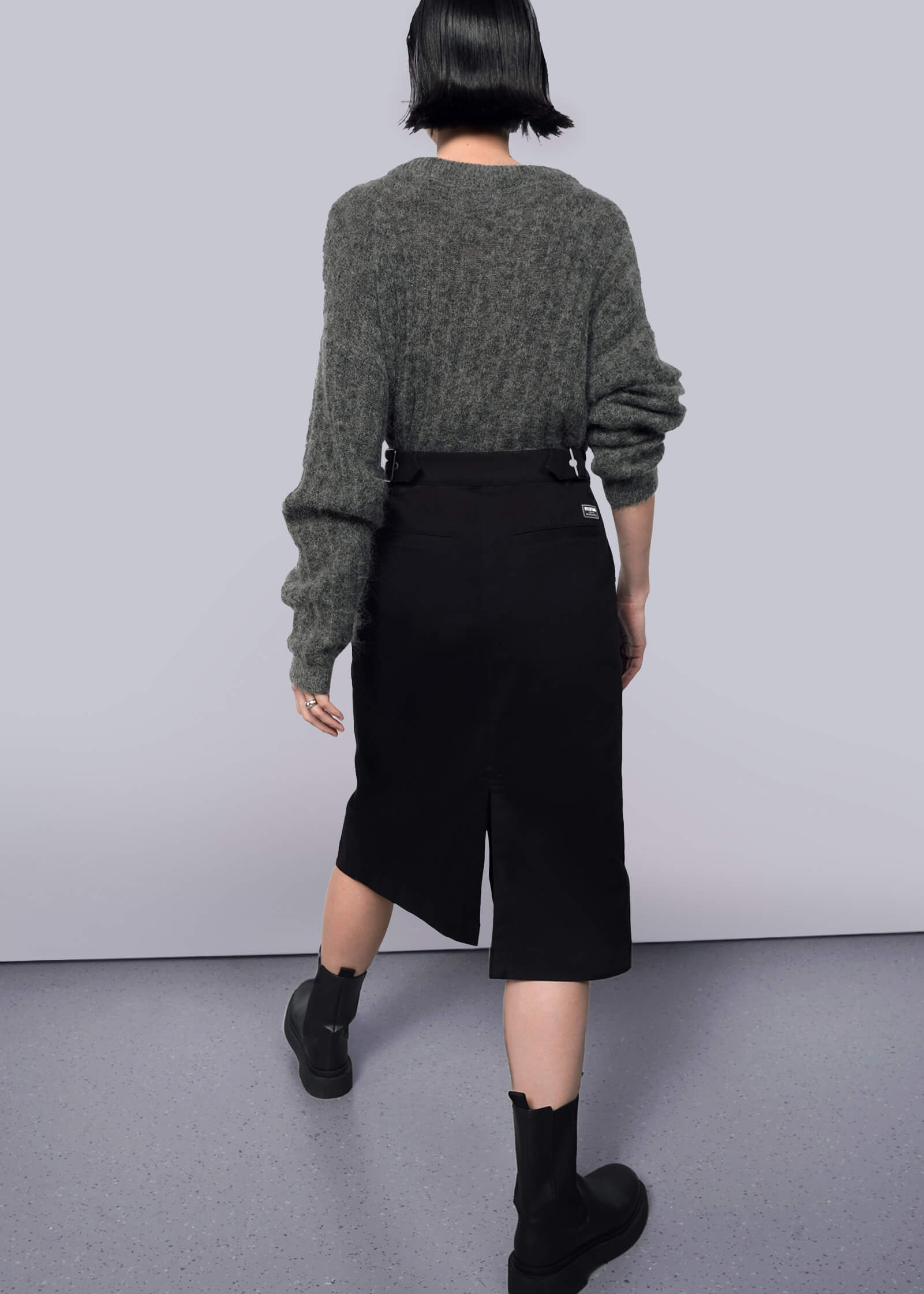S= A person with short dark hair faces away, wearing a loose gray sweater with The Essential Utility Wrap Skirt—black, midi-length with back slit and adjustable waistband—and black ankle boots, against a plain white background.