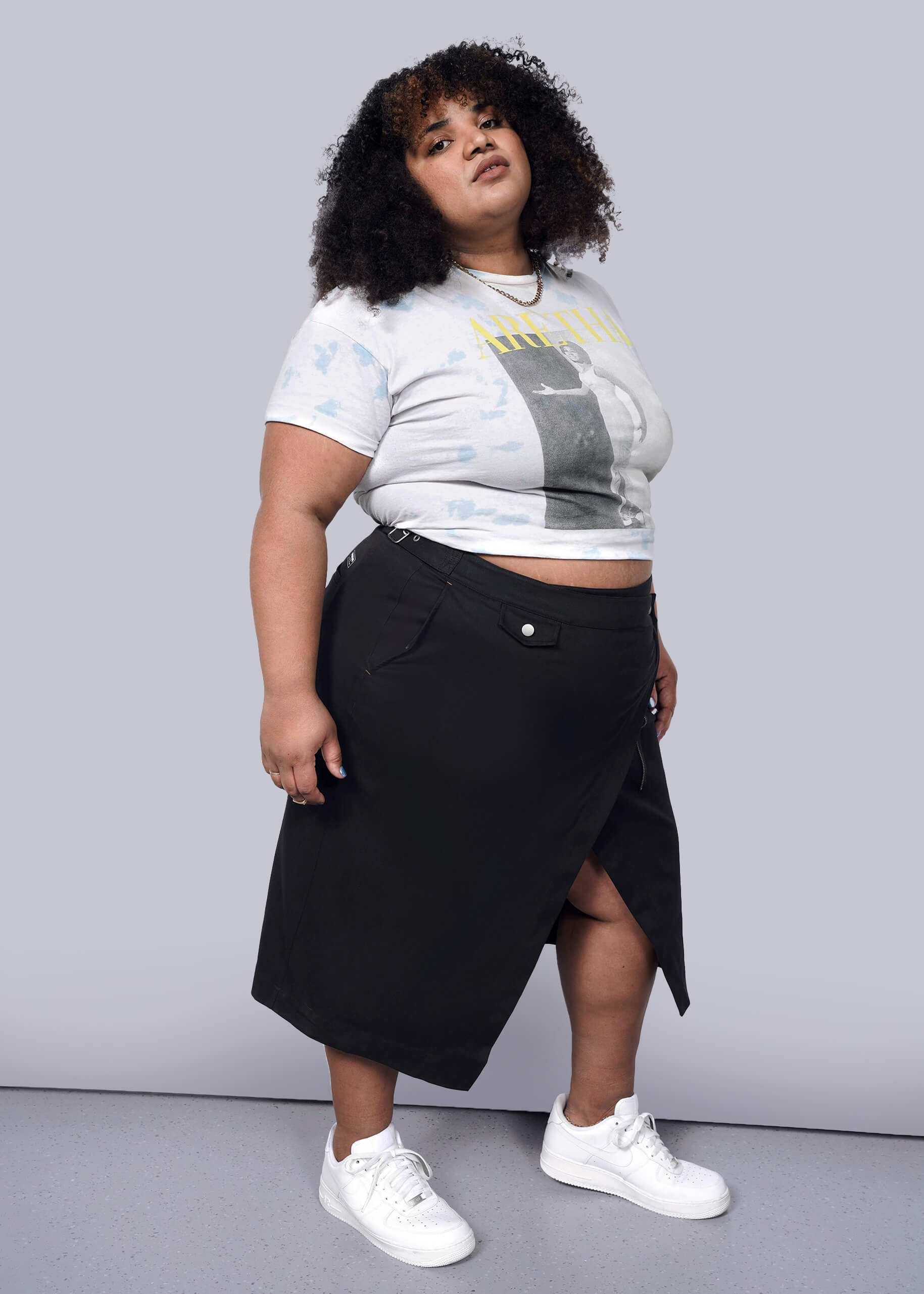 1X=A person with curly hair wears a graphic crop top, The Essential Utility Wrap Skirt in black with an asymmetrical waist detail, and white sneakers, standing confidently against a plain white background.