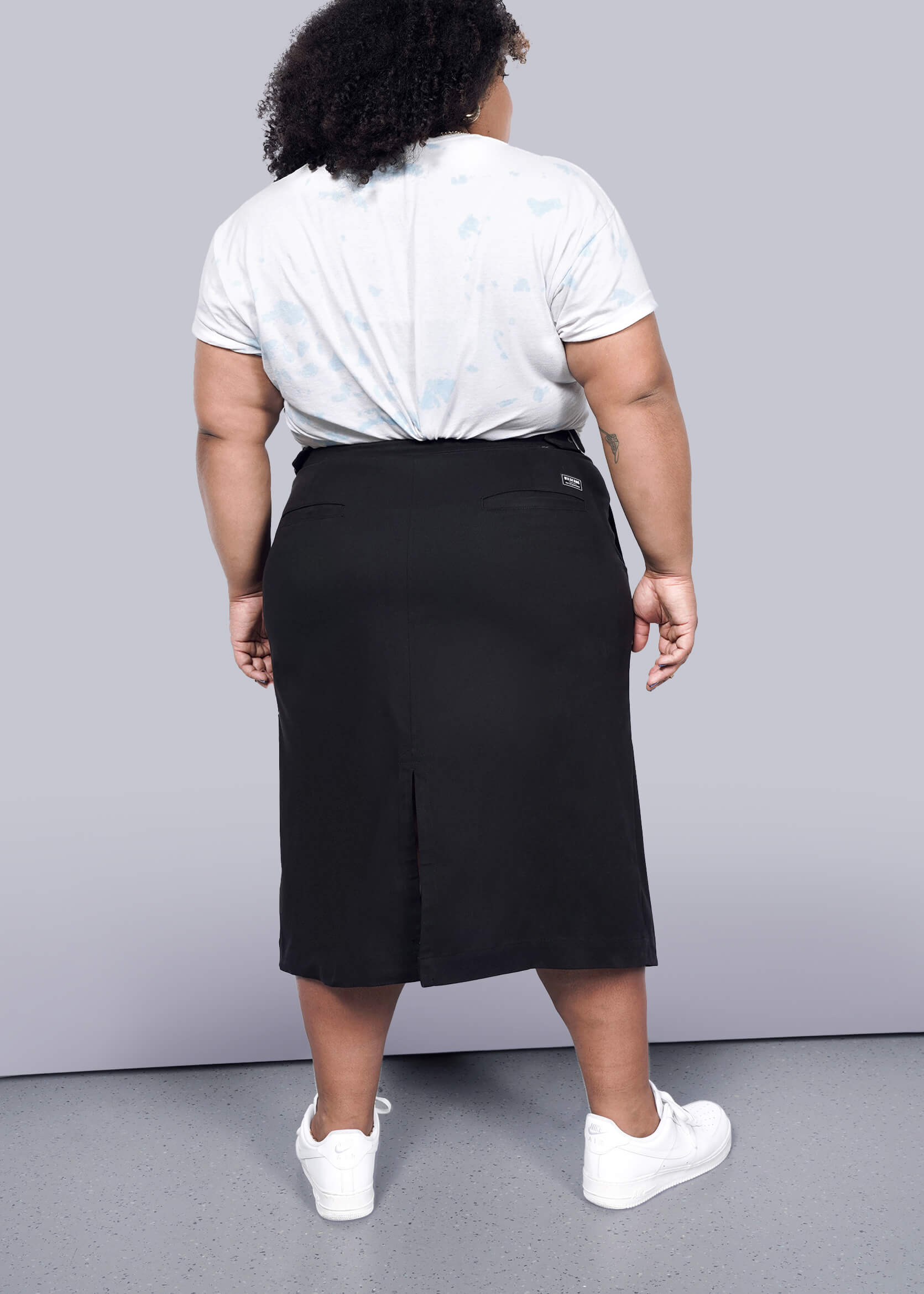 1X= A person with curly hair faces away, wearing a light t-shirt, The Essential Utility Wrap Skirt in black with an asymmetrical waist, and white sneakers against a white background.