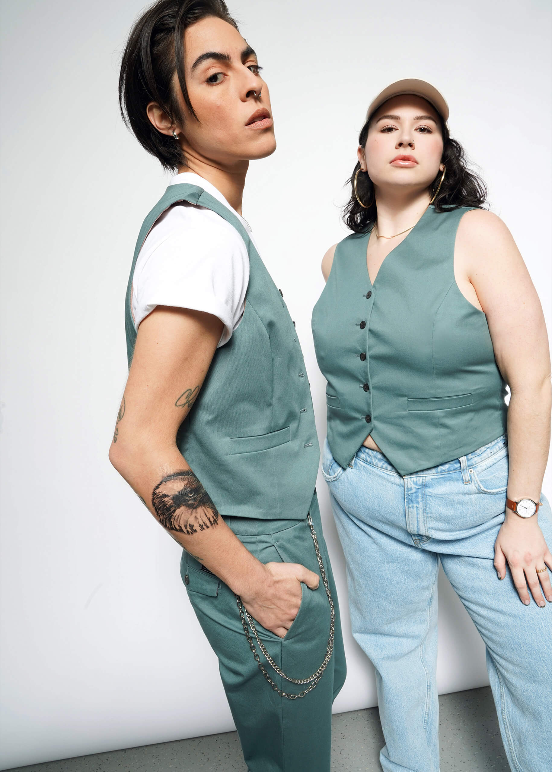 S=Two people pose confidently. One wears The Essential Vest in green with matching pants, white T-shirt, and chain; the other styles The Essential Vest in green with 5-button closure, light blue jeans, beige cap, and watch.
