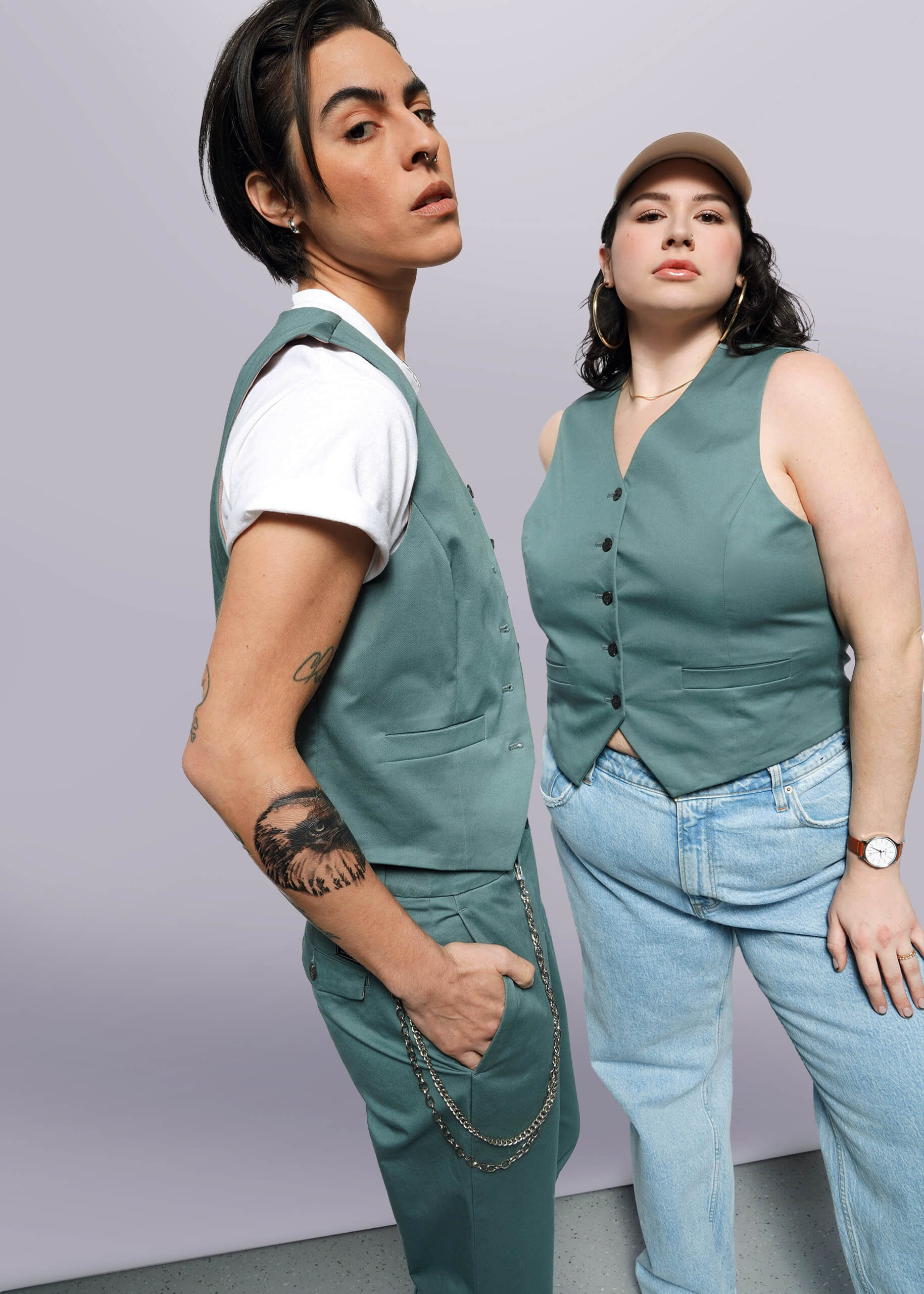S=Two people pose confidently. One wears The Essential Vest in green with matching pants, white T-shirt, and chain; the other styles The Essential Vest in green with 5-button closure, light blue jeans, beige cap, and watch.