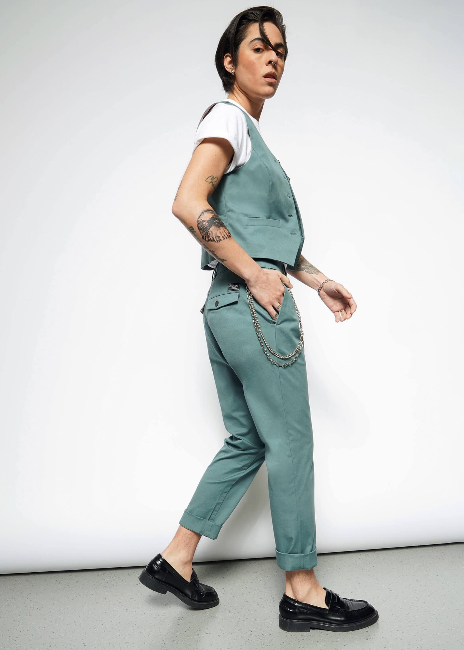 S=A tattooed person in a white t-shirt, The Essential Vest—tailored fit stretch cotton with 5 buttons—teal pants with a chain, and black loafers walks confidently against a plain white background.