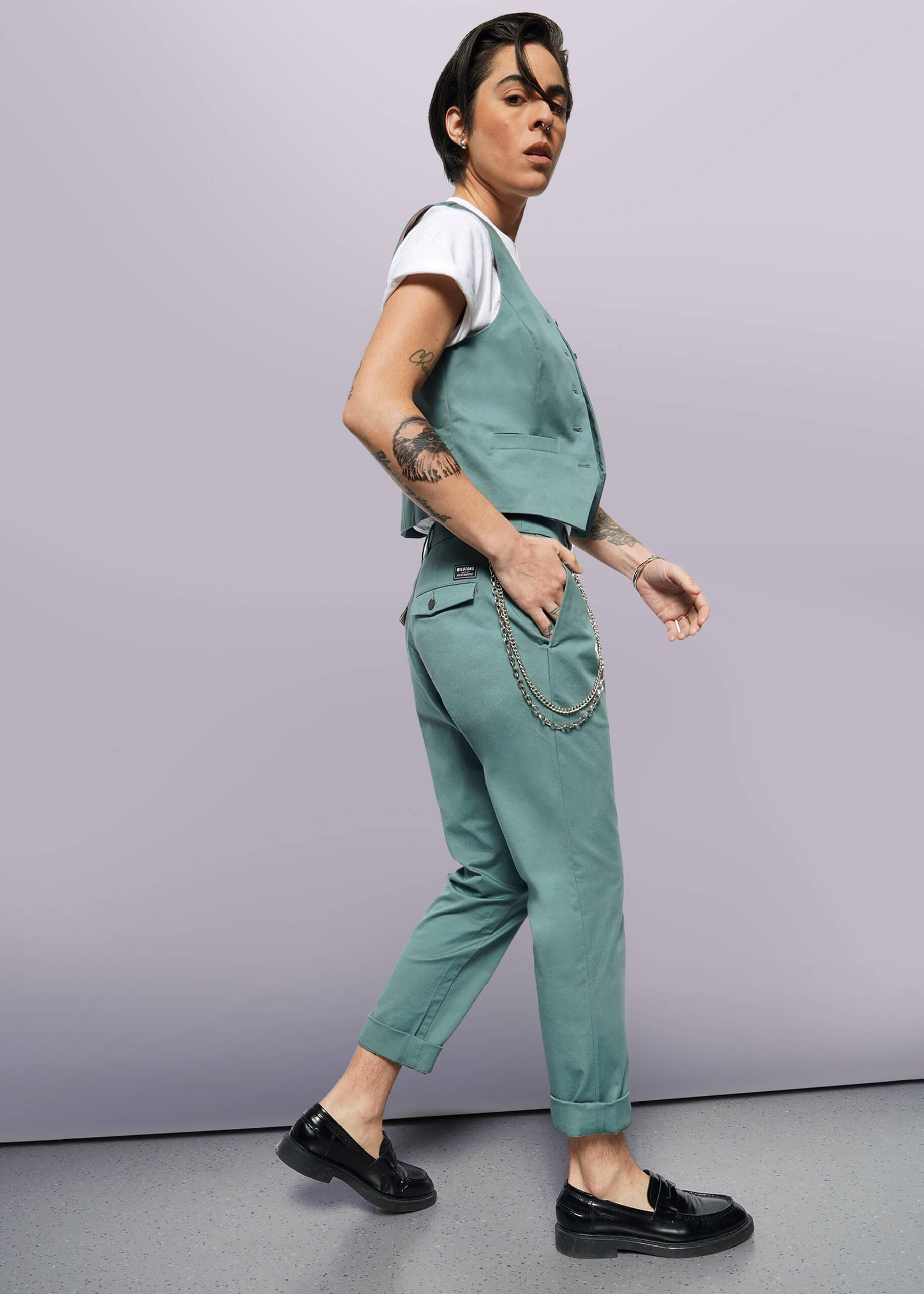 S=A tattooed person in a white t-shirt, The Essential Vest—tailored fit stretch cotton with 5 buttons—teal pants with a chain, and black loafers walks confidently against a plain white background.