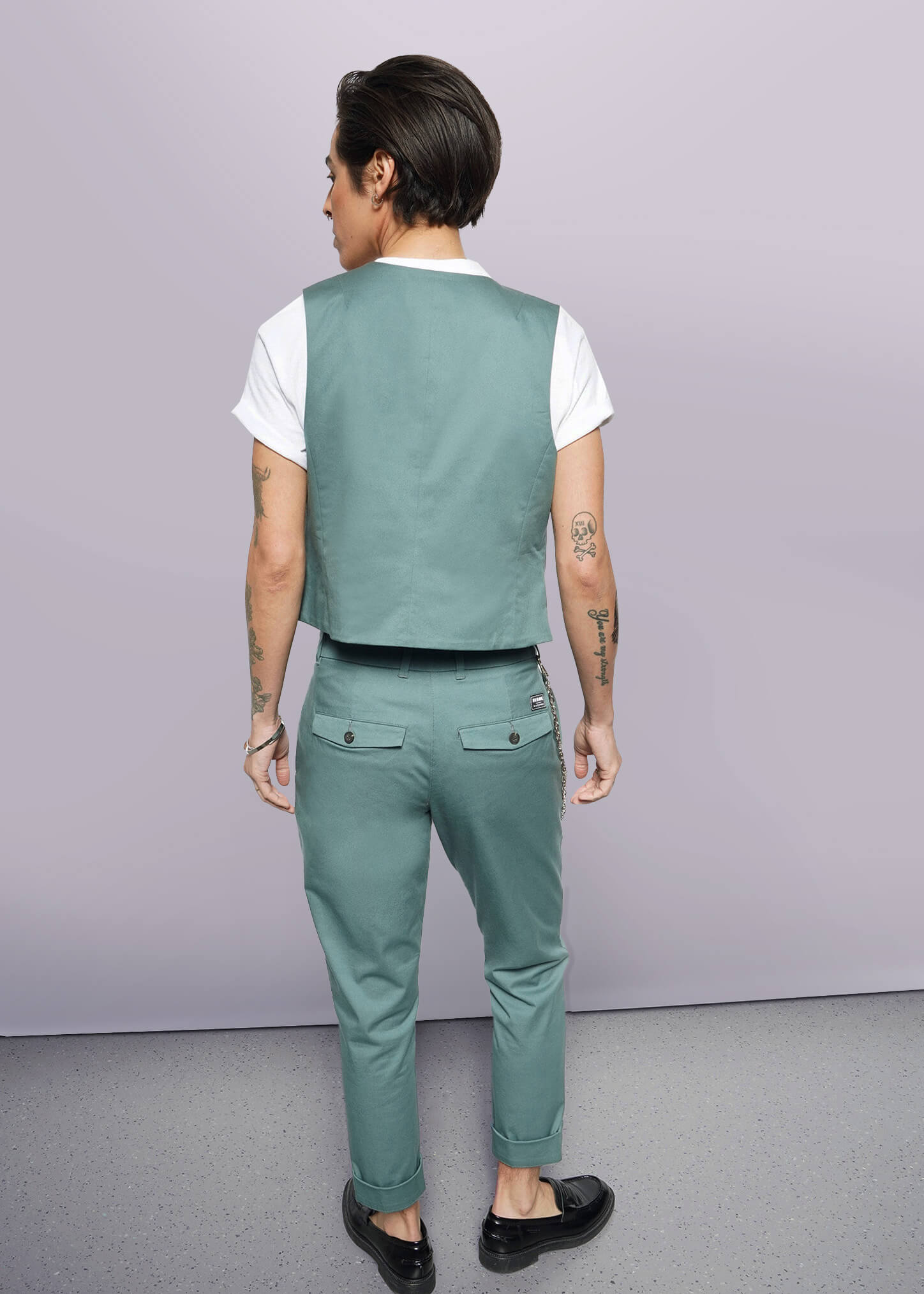 S=A person with short dark hair, wearing The Essential Vest—tailored fit, stretch cotton, 5-button closure—with matching pants, white T-shirt, and black shoes stands facing away from the camera, displaying arm tattoos against a plain white background.