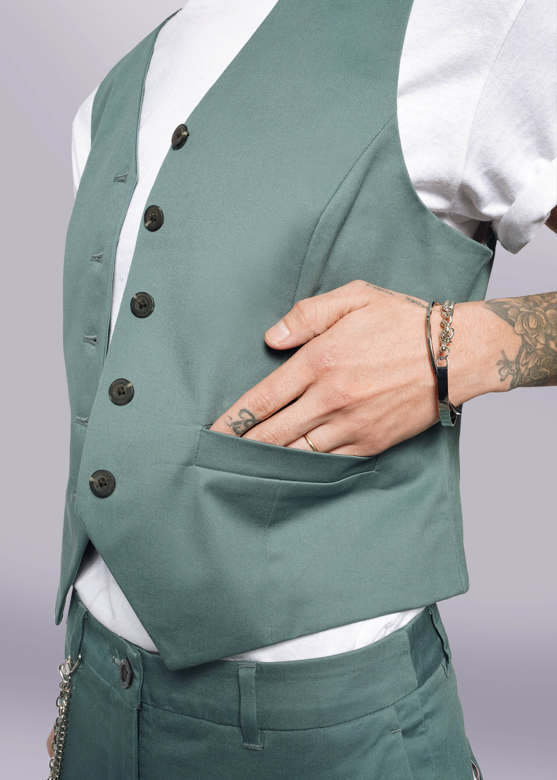 S= Wearing The Essential Vest in sage green stretch cotton with a tailored fit and 5-button closure, paired with matching pants and a white T-shirt. A tattooed hand rests in the vest pocket, displaying a bracelet and ring; cropped torso shot.