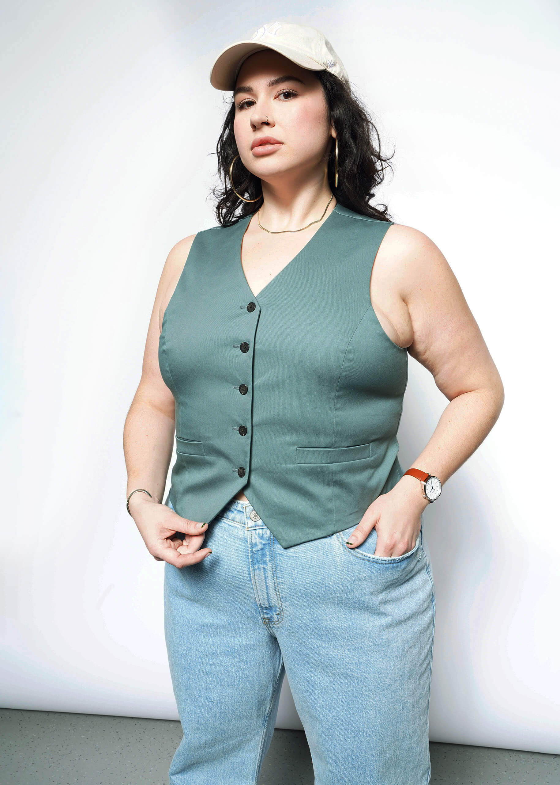 1X= A woman poses confidently against a light background, wearing The Essential Vest—a light tan, stretch cotton vest with 5-button closure—paired with light blue jeans and a wristwatch, one hand in her pocket and the other holding her vest.