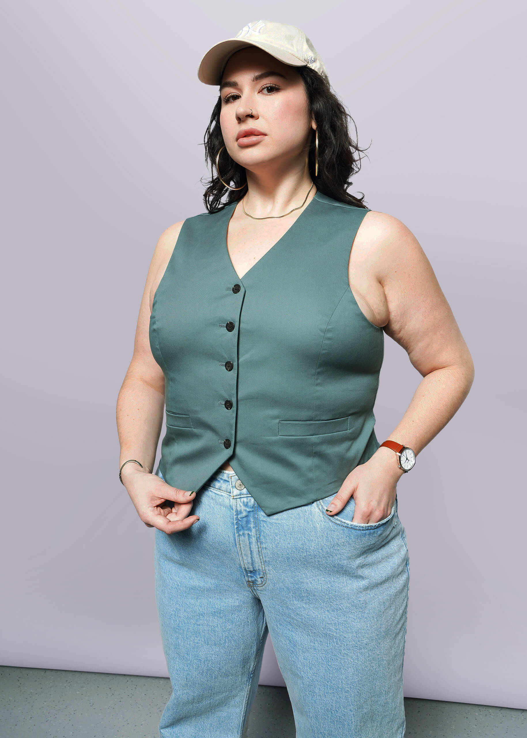 1X= A woman poses confidently against a light background, wearing The Essential Vest—a light tan, stretch cotton vest with 5-button closure—paired with light blue jeans and a wristwatch, one hand in her pocket and the other holding her vest.