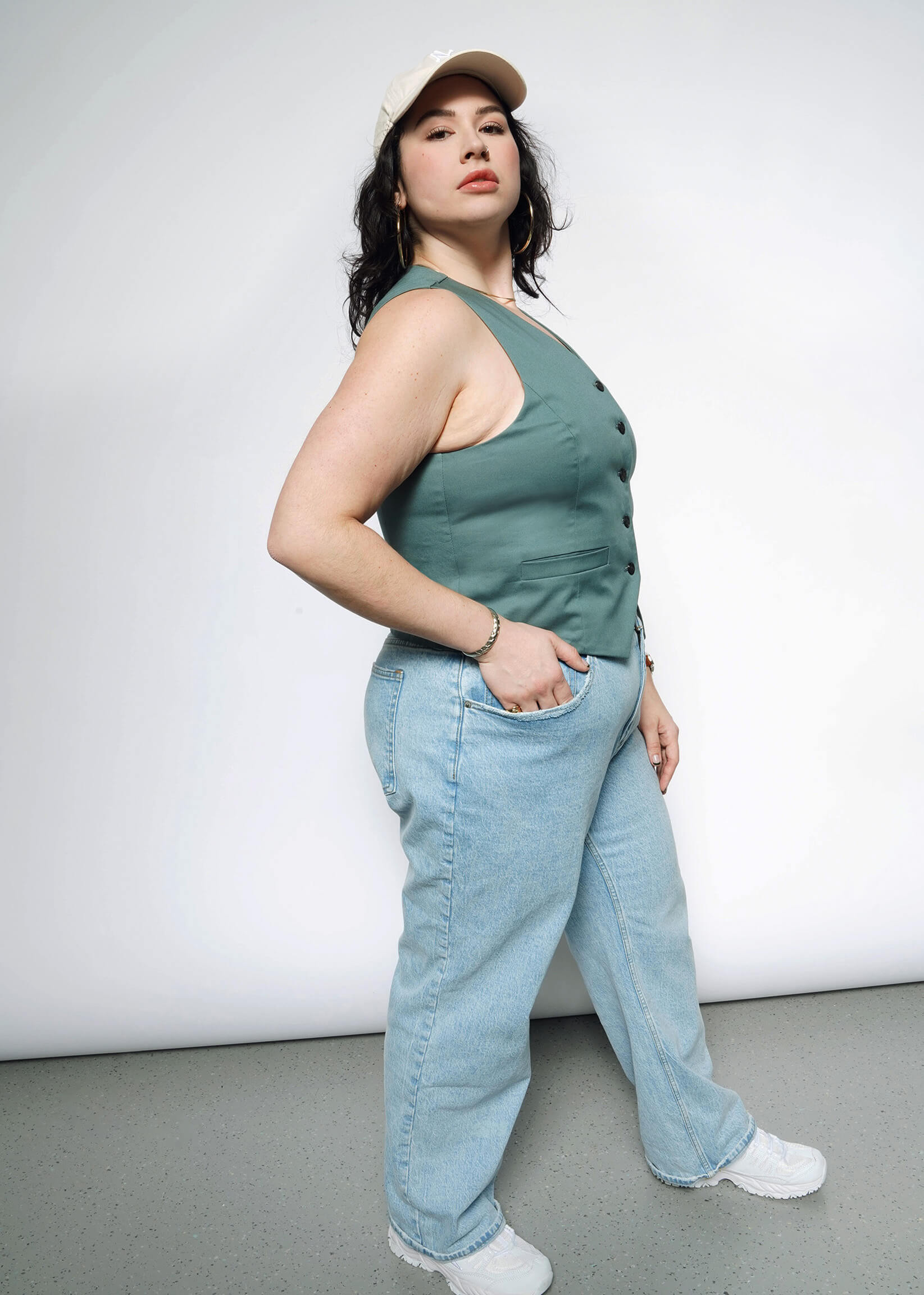 1X=A woman confidently showcases The Essential Vest, a tailored stretch cotton piece with a 5-button closure, paired with a beige cap, light blue jeans, and white sneakers, standing against a plain white background.