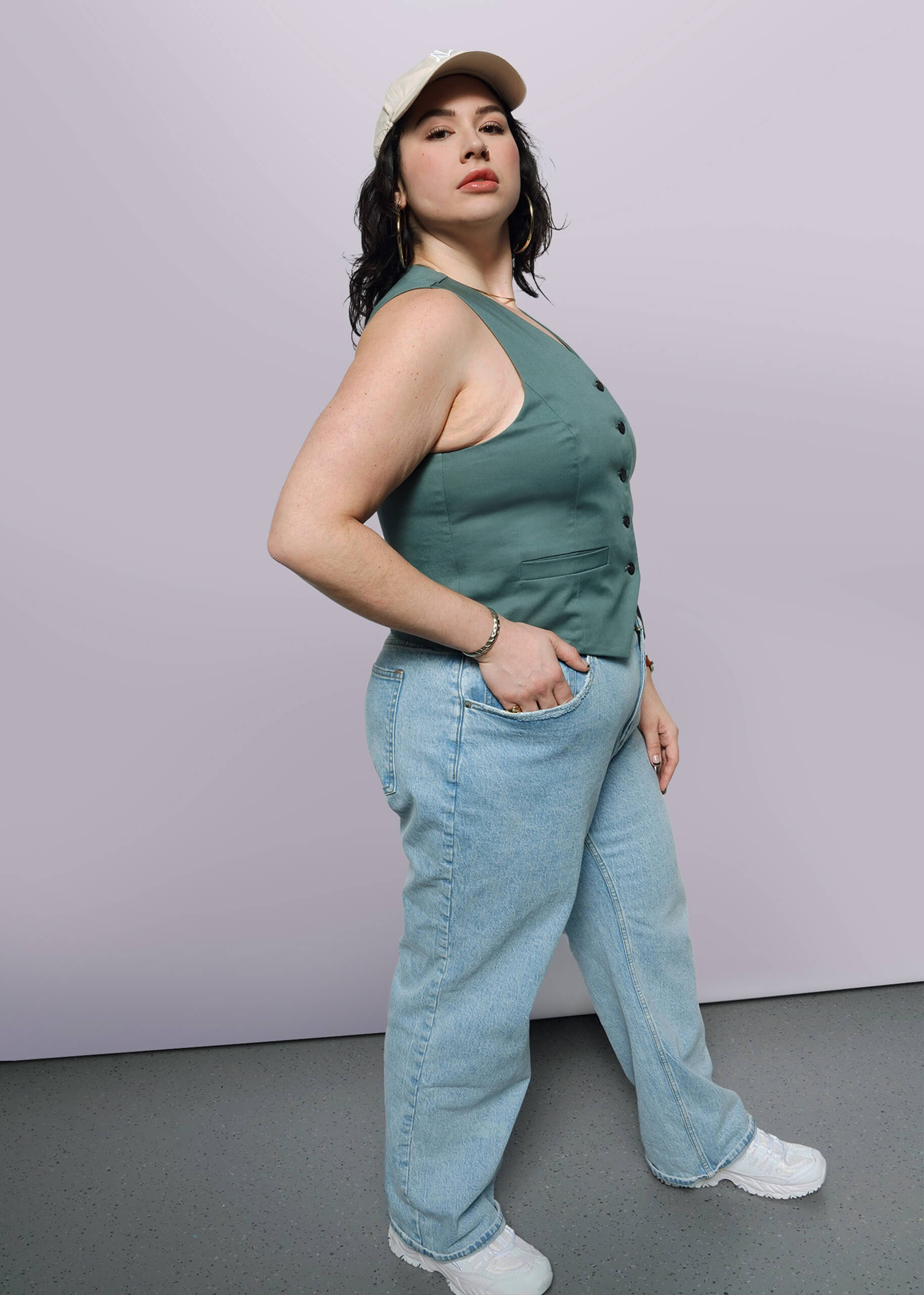 1X=A woman confidently showcases The Essential Vest, a tailored stretch cotton piece with a 5-button closure, paired with a beige cap, light blue jeans, and white sneakers, standing against a plain white background.