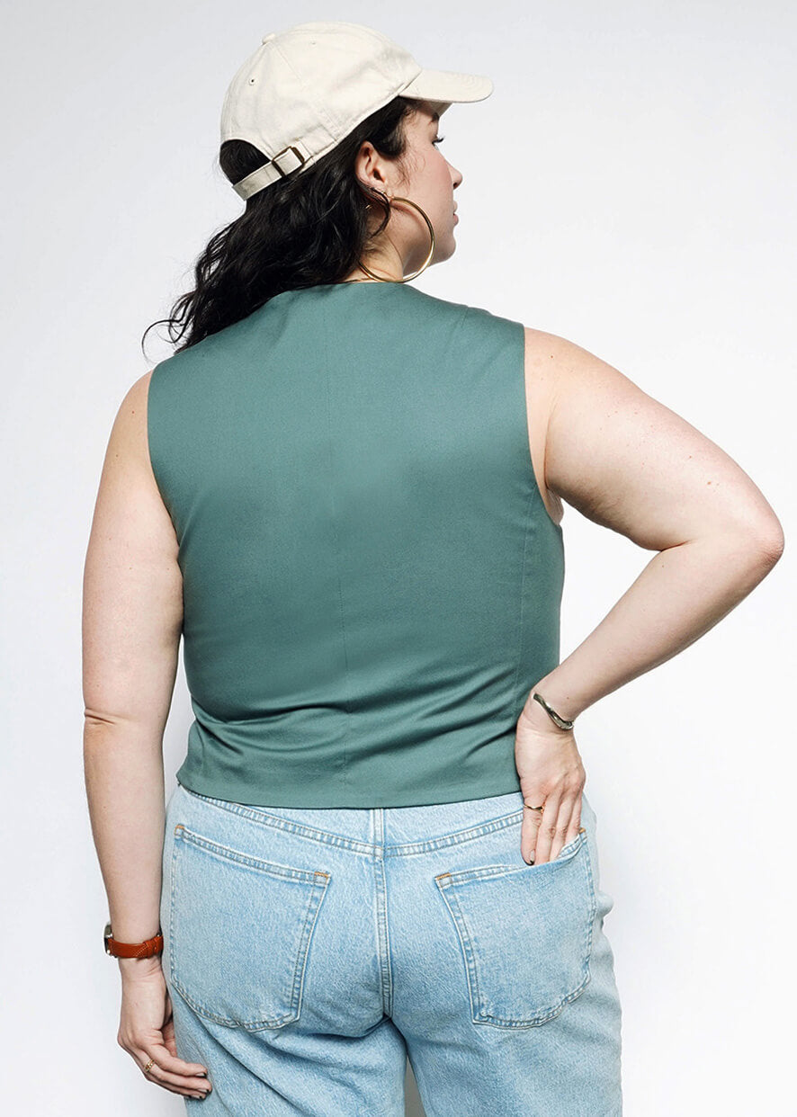 1X= Wearing The Essential Vest with a tailored fit, a light beige cap, and light blue jeans, a person stands with their back to the camera—one hand on their hip and the other by their side.