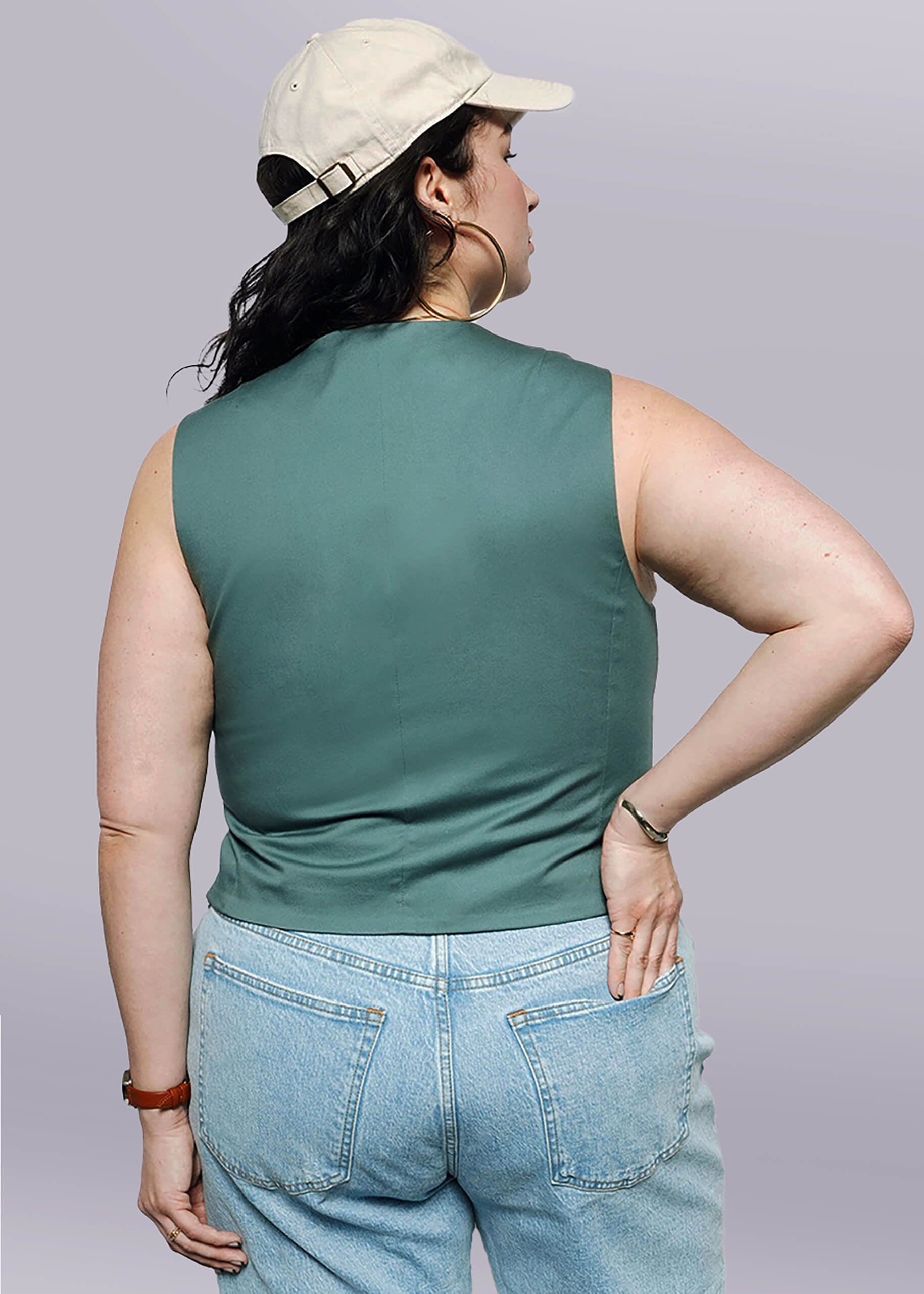 1X= Wearing The Essential Vest with a tailored fit, a light beige cap, and light blue jeans, a person stands with their back to the camera—one hand on their hip and the other by their side.