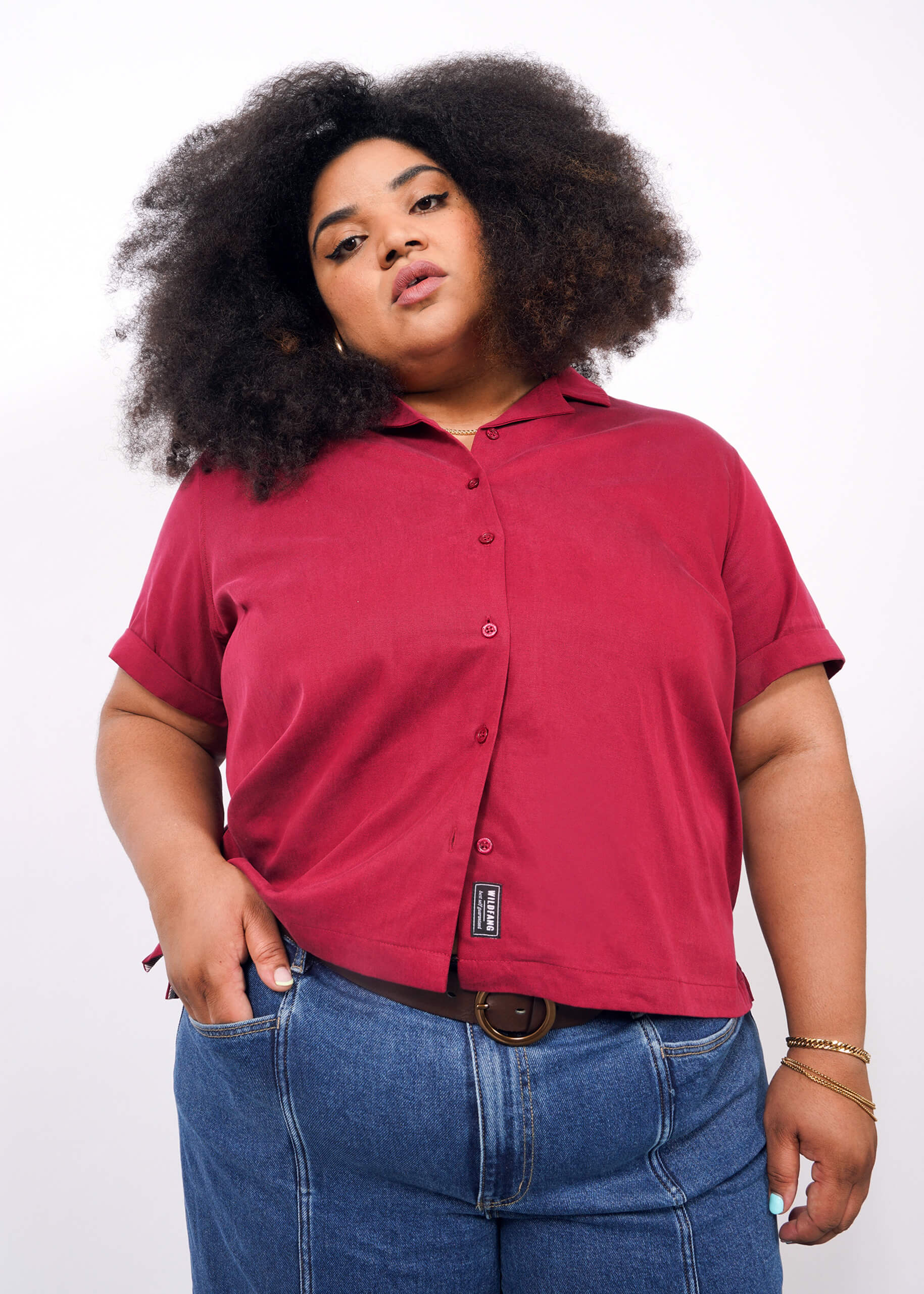 1X=A person with voluminous curly hair wears The Everyday Camp Button Up in dark red, featuring a relaxed fit and ultra-soft fabric, paired with blue jeans. They stand against a plain white background, looking confidently at the camera.