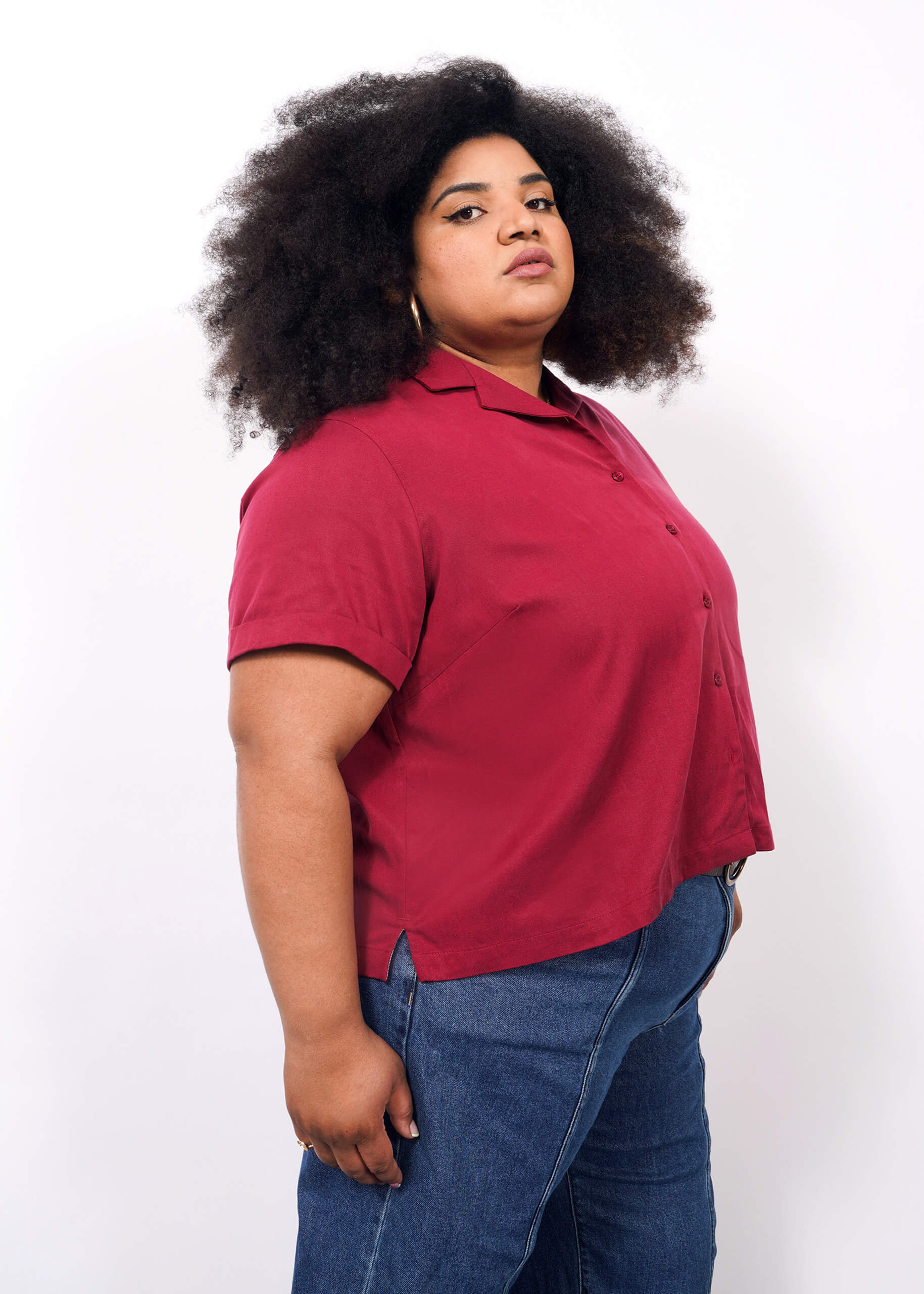 1X=A person with curly, voluminous hair wears The Everyday Camp Button Up—a relaxed fit red camp collar shirt in ultra-soft fabric—and blue jeans, standing in profile against a plain white background, looking confidently at the camera.