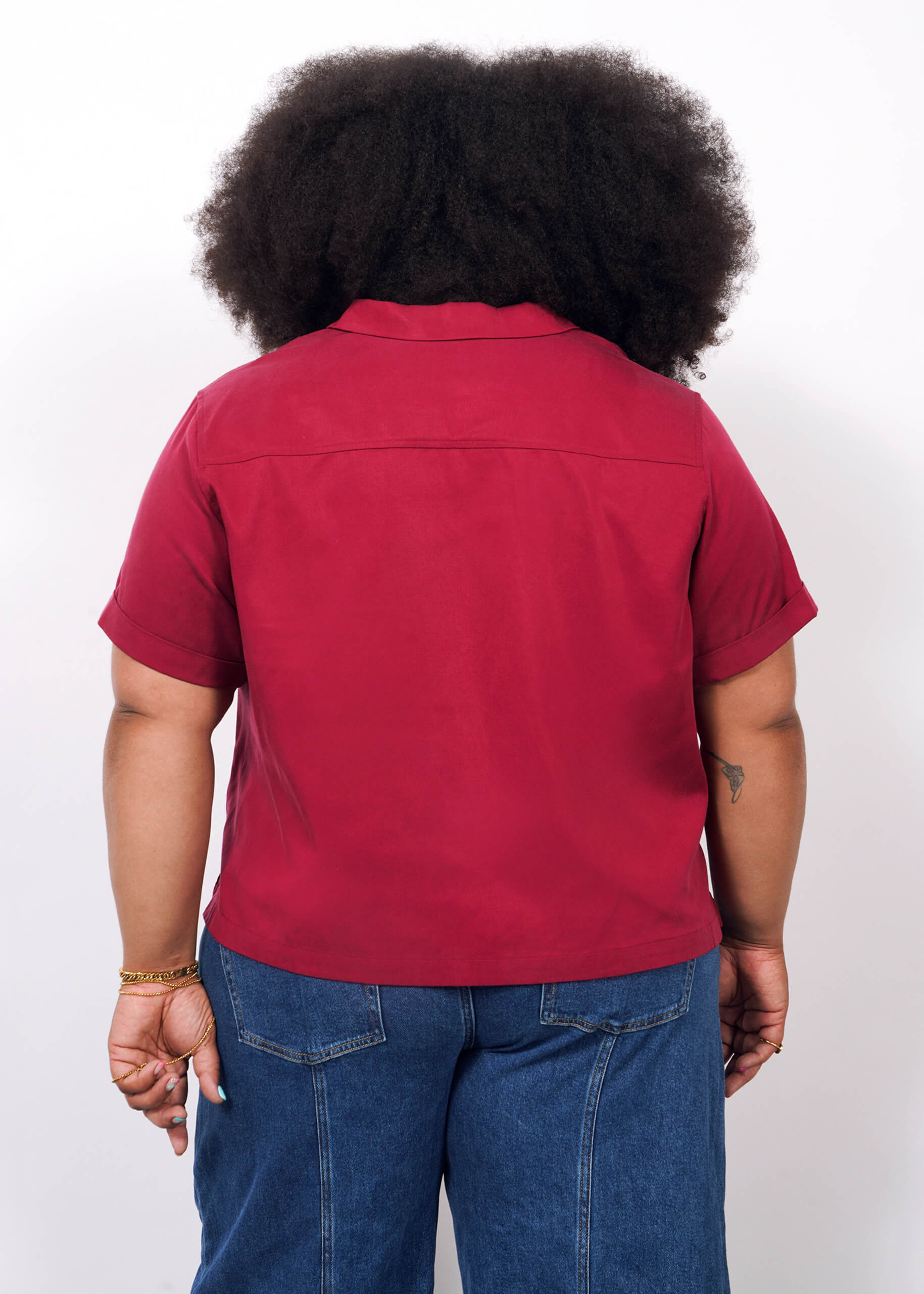 1X=A person with curly hair stands with their back to the camera, wearing The Everyday Camp Button Up in red and blue jeans, set against a plain white background.