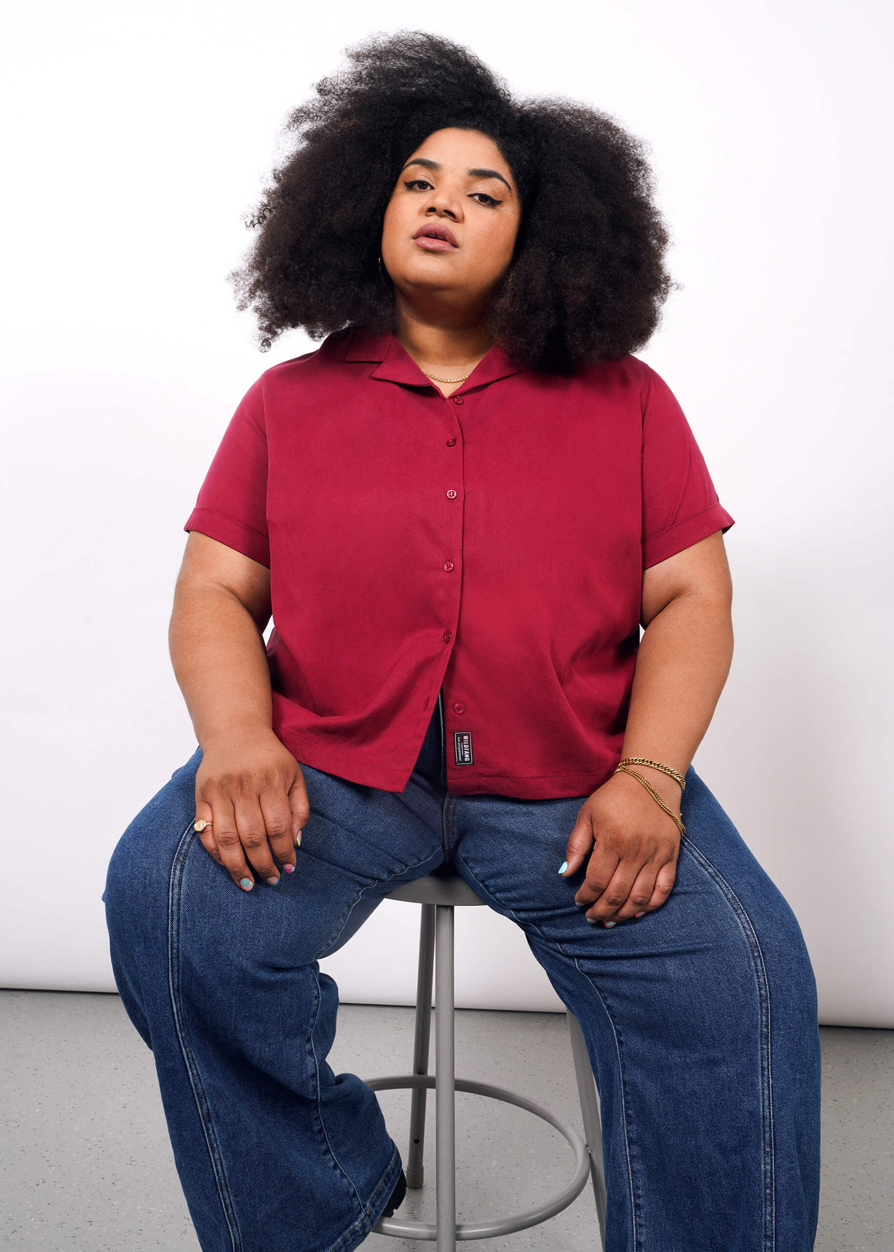 1X= A woman with voluminous curly hair sits on a stool against a white background, wearing The Everyday Camp Button Up in red and wide-leg blue jeans. She looks confidently at the camera, her hands resting on her thighs.