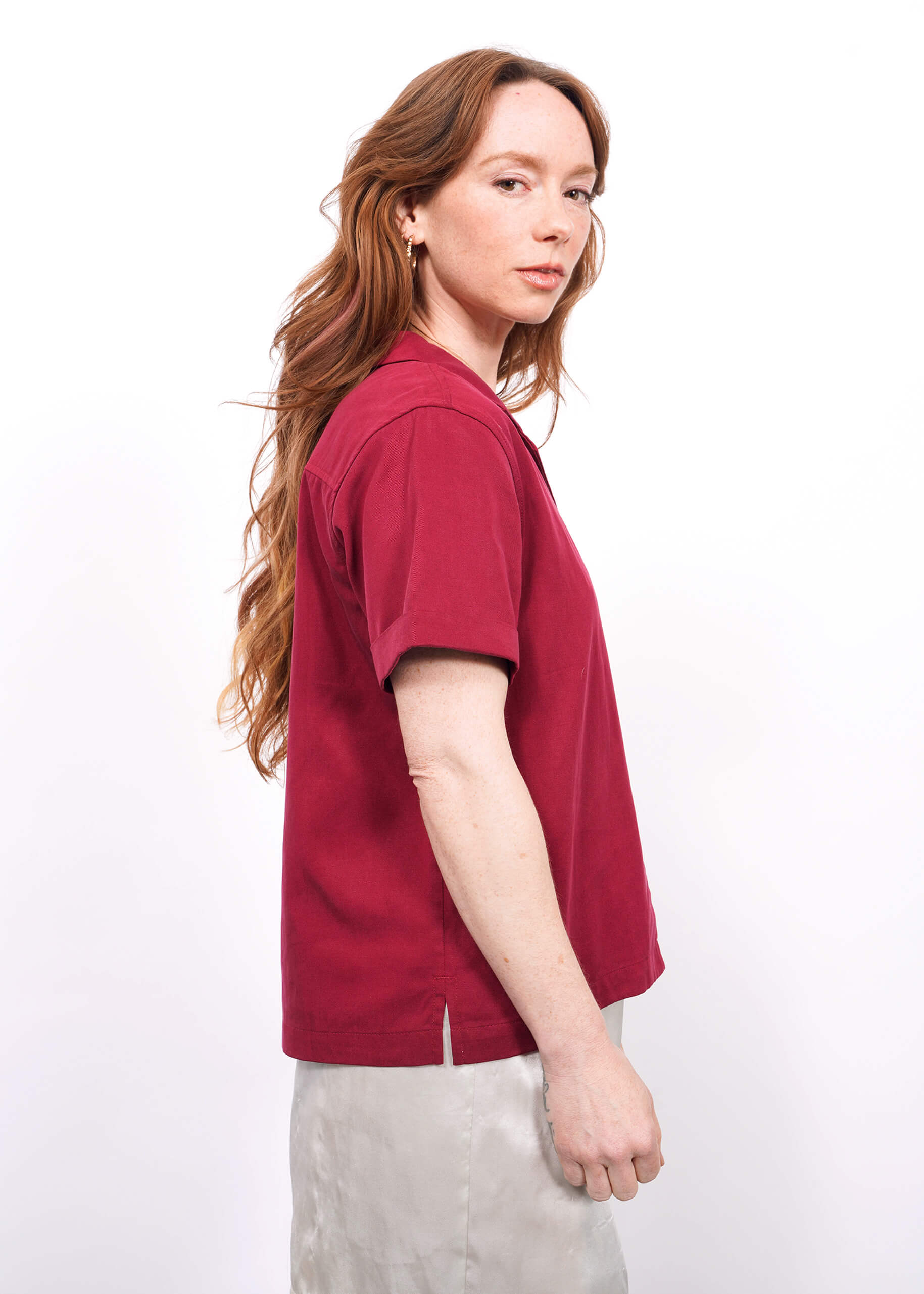 S= A woman with long, wavy auburn hair stands in profile, wearing The Everyday Camp Button Up in dark pink and a light-colored skirt, against a plain white background.