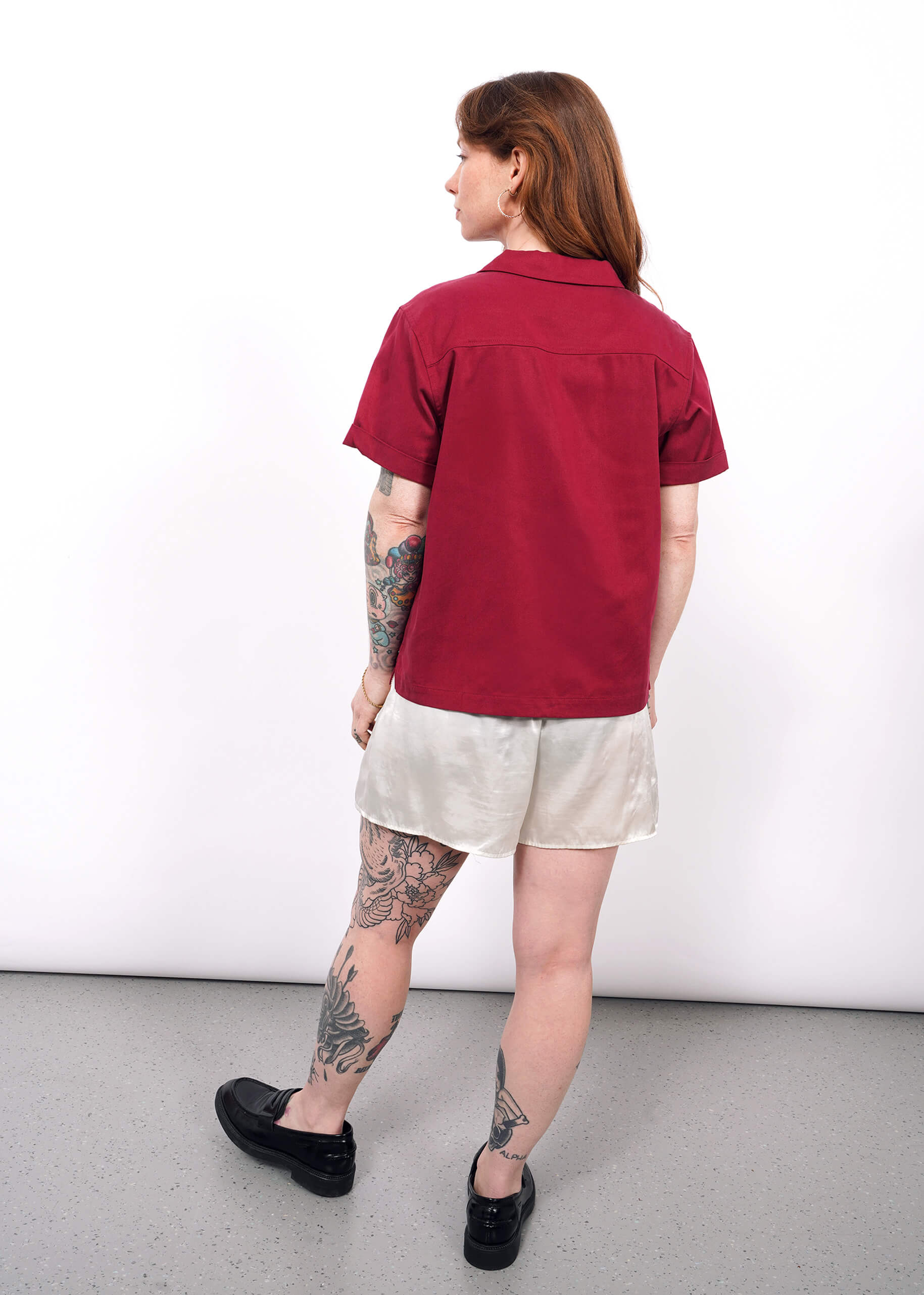 S=A woman with tattoos stands facing away in The Everyday Camp Button Up—relaxed fit, maroon color—paired with light shorts and black shoes, against a plain white background.