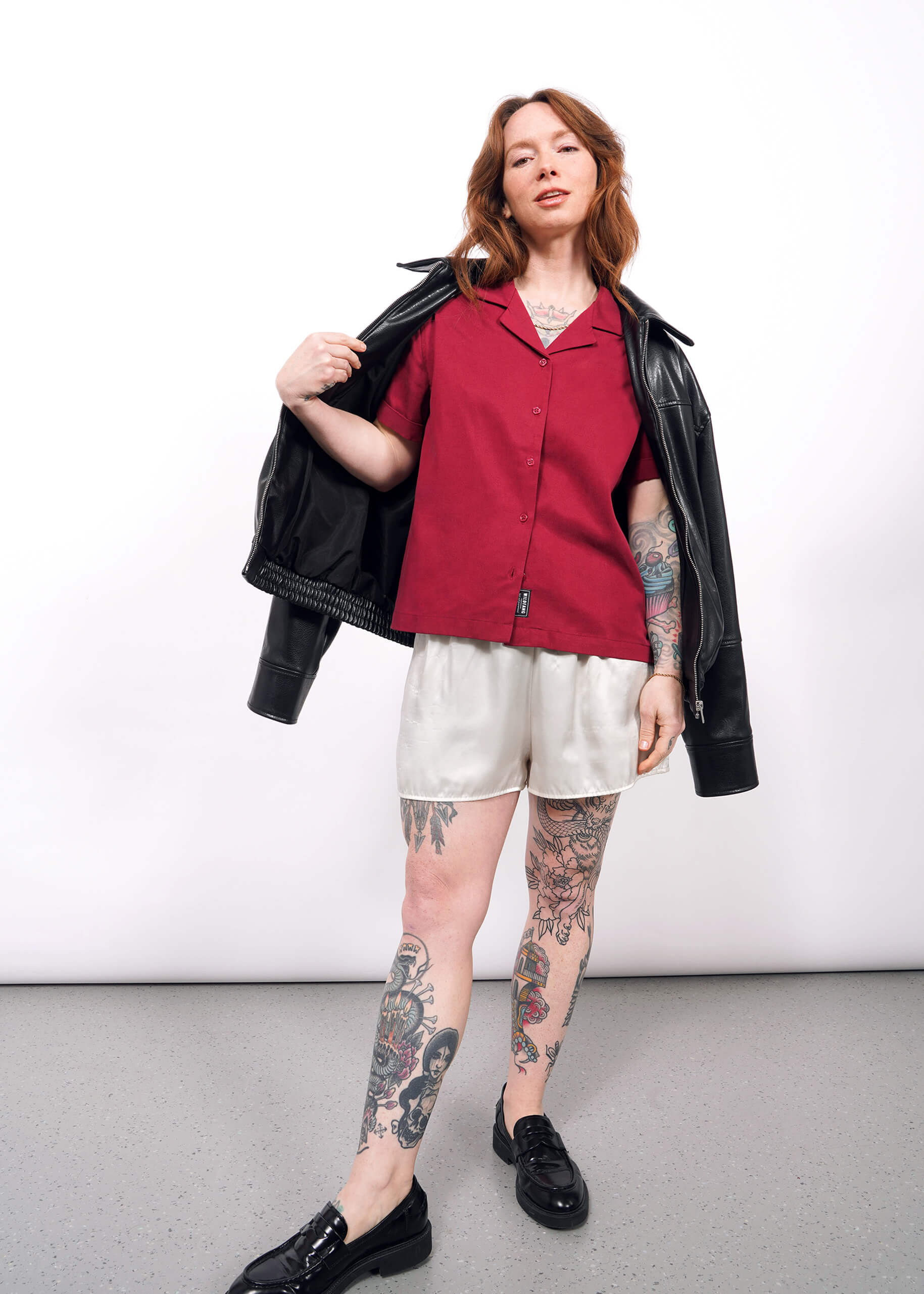 S=A tattooed woman stands against a white background wearing The Everyday Camp Button Up in red, white satin shorts, black shoes, and a black leather jacket draped over her shoulders.